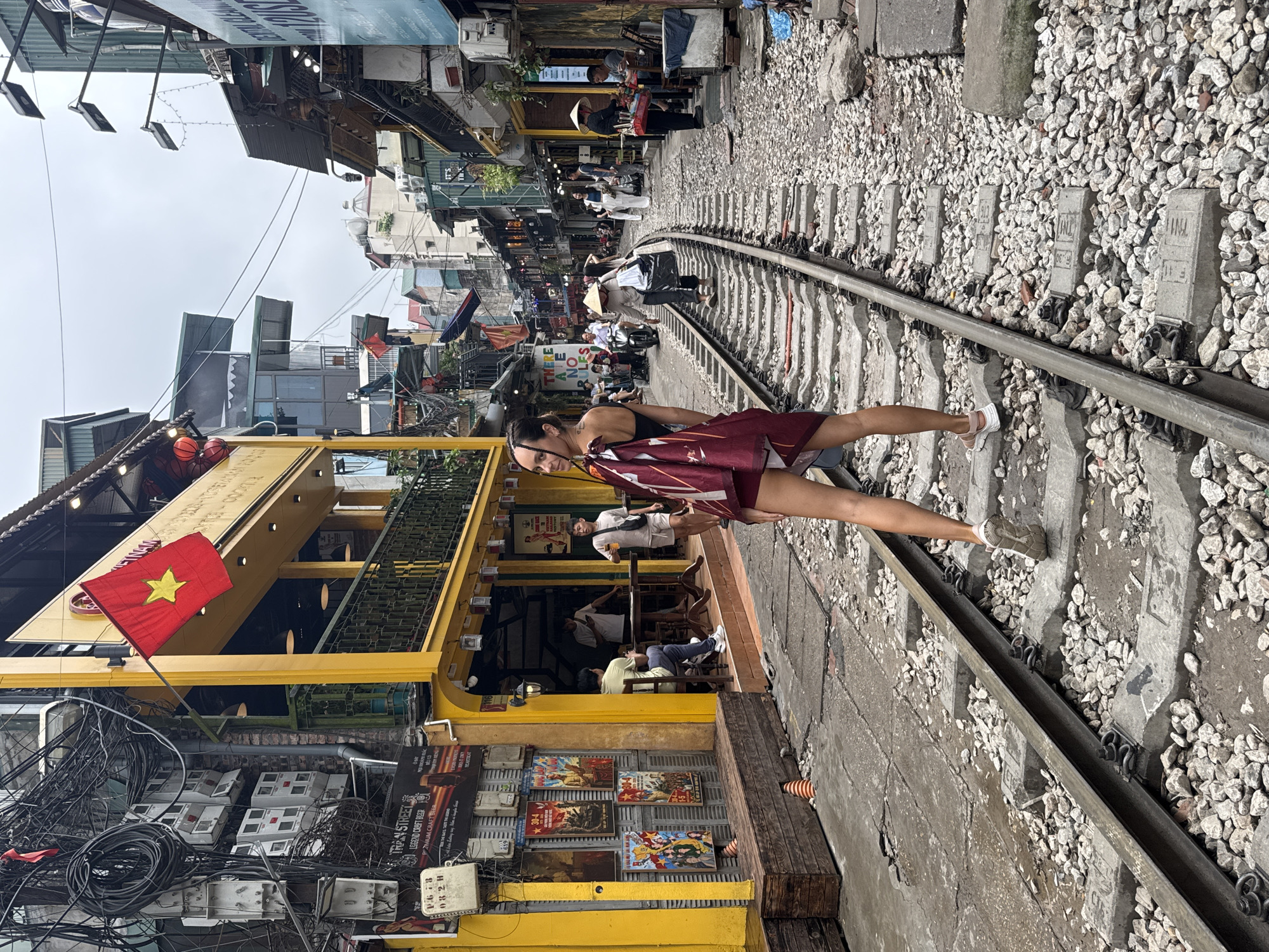 The image captures a bustling scene on a railway track running through the heart of Hà Nội, Vietnam. A woman, standing on the track, is positioned in the foreground. The railway line stretches into the distance, interspersed with gravel and concrete sleepers. Overhead, a chaotic network of tangled electrical cables hangs above the street. The buildings lining the track exhibit a traditional Vietnamese architectural style. A Vietnamese flag hangs prominently from a yellow building that houses a bar or restaurant. Tables and chairs are arranged indoors, with several people seated. Posters adorn the exterior of the building. The image encapsulates the unique character of Hà Nội, where daily life coexists with the active railway line.
