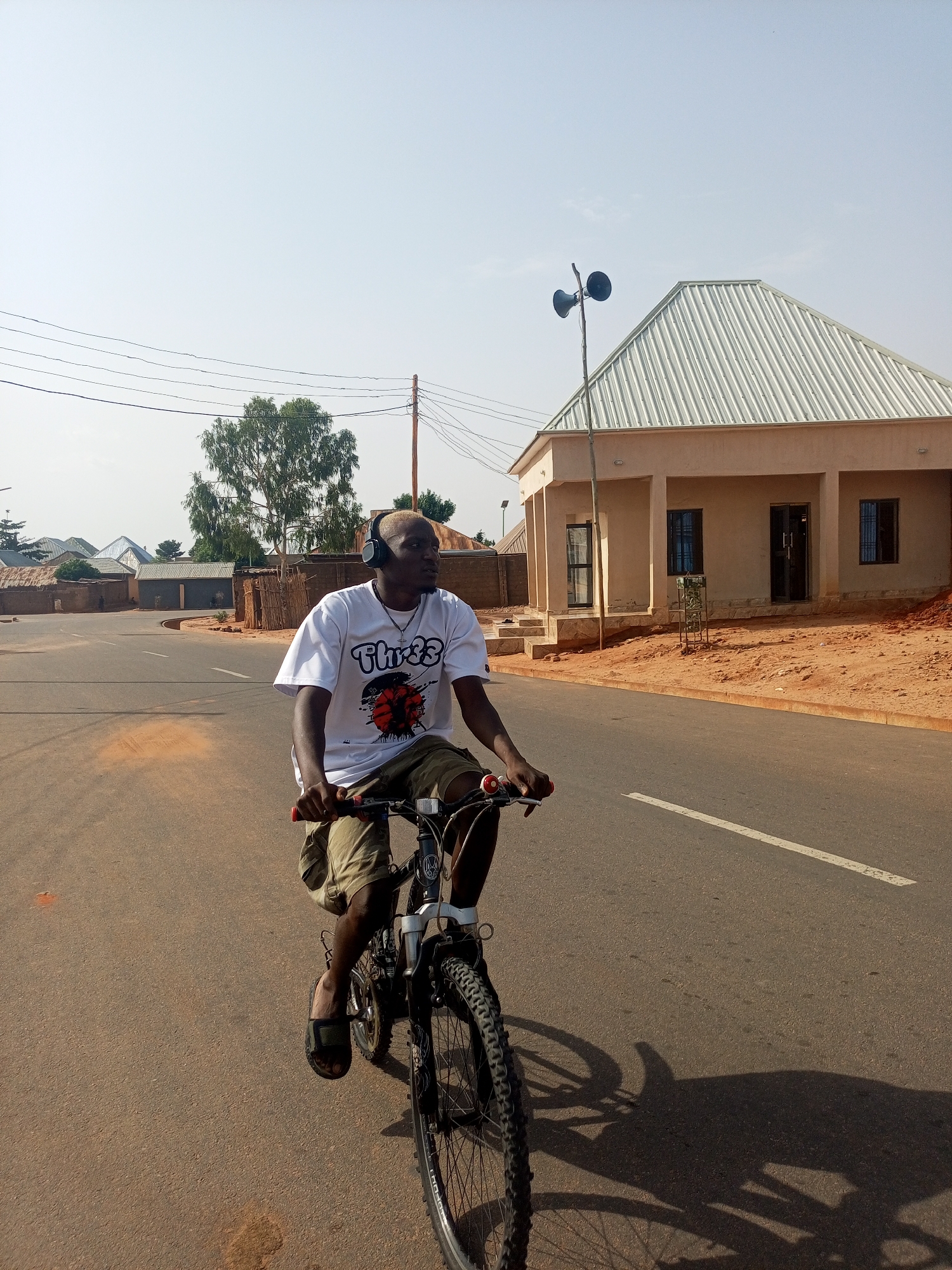 A young Black man rides a mountain bike on a paved road in Tempure, Nigeria. He wears headphones, a white t-shirt, khaki shorts, and sandals. He is the sole person shown. The road is lined with modest buildings and trees; a building with loudspeakers attached to a pole is on the right. The sky is bright.