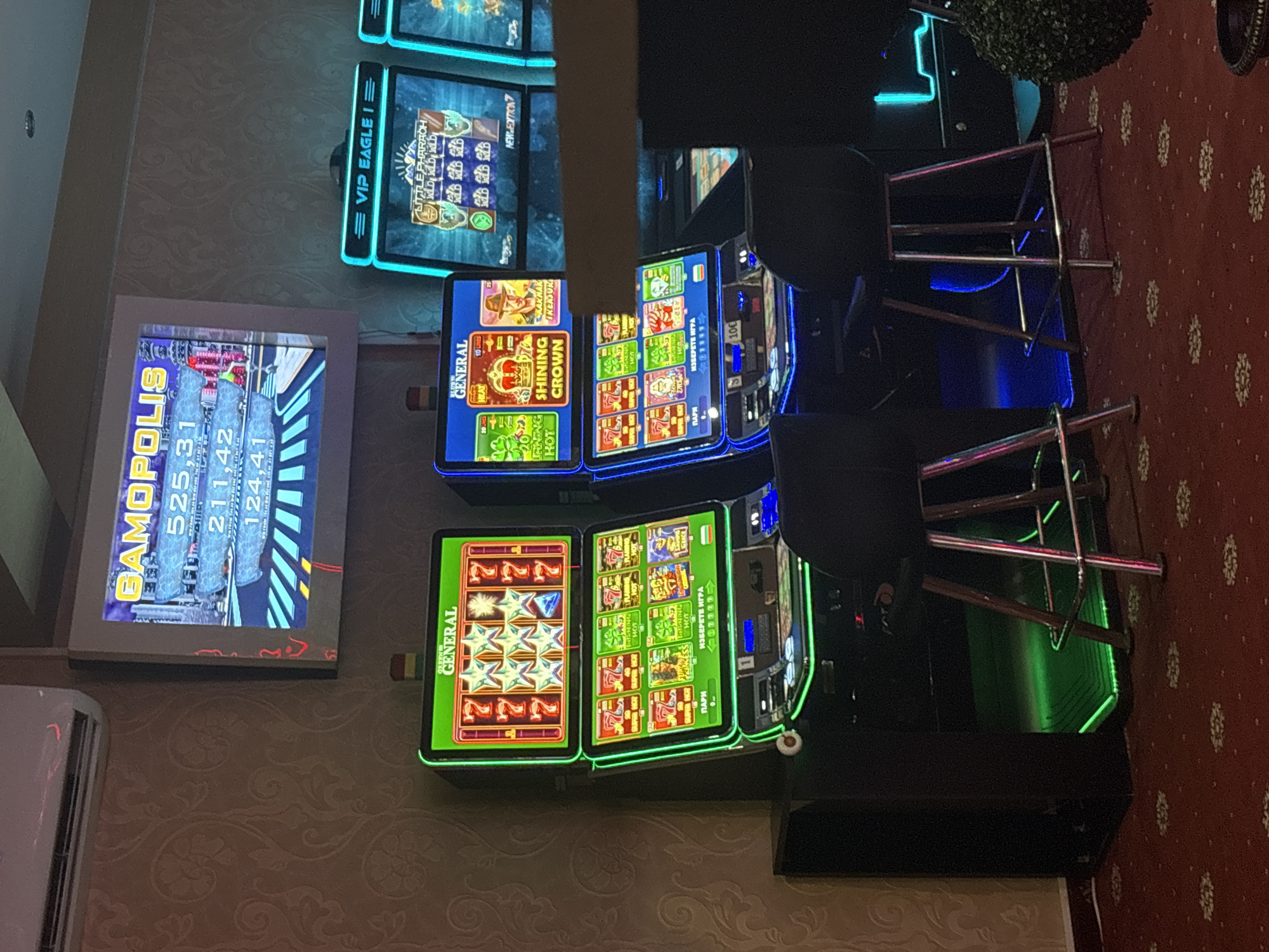 This is a photograph of a dimly lit casino interior, specifically a section with slot machines. In the foreground, two bar stools with metallic legs and black seats are positioned in front of slot machines. The slot machines are illuminated with bright green and blue lights. The machine on the left is a "GENERAL" branded slot machine with a display showing various symbols, including lucky sevens and stars, as well as game titles like "FURRY PRINCESS". The machine on the right also features a "GENERAL" branded screen with game titles like "SHINING CROWN" and "RAKHAR TREASURE", accompanied by vibrant graphics. Further to the right, another slot machine with a blue neon light frame and a "VIP EAGLE" title is visible, displaying "LITTLE PHARAOH" game graphics. In the background, a large screen mounted on the wall displays colorful graphics and text, including "GAMOPOLIS" and numerical values, likely indicating game scores or jackpots. The walls are adorned with a textured wallpaper featuring a damask pattern. The floor is covered with a red carpet with a scattered floral design. An air conditioning unit is visible on the upper left wall. The overall ambiance suggests a gaming establishment in Karlovo, Bulgaria.
