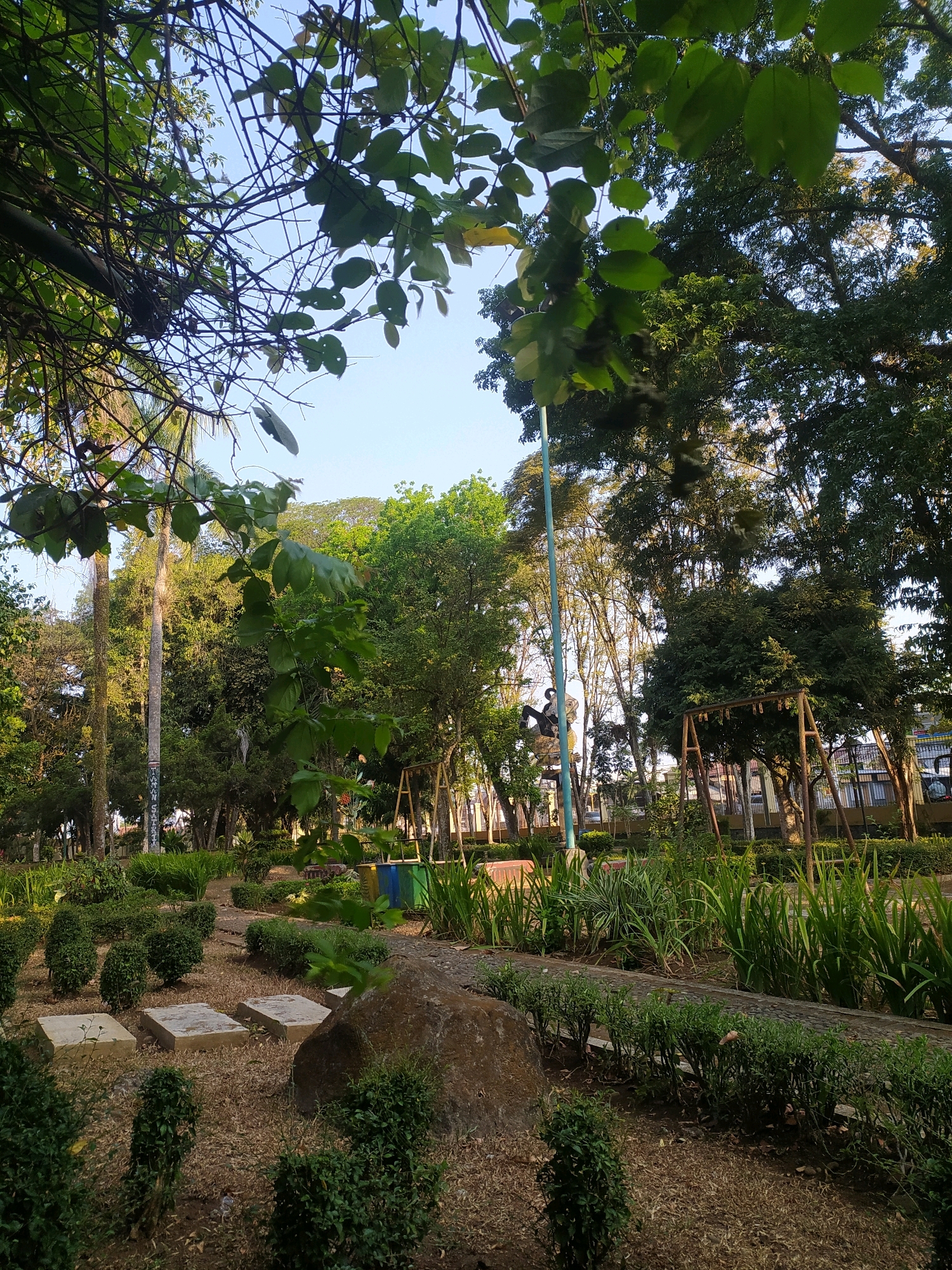 The image depicts a park-like setting with a clear sky. In the foreground, there are several square stepping stones placed on dry earth, surrounded by low-lying green bushes. A large, rough-textured rock is positioned to the right of the stepping stones. Beyond this, a paved path is visible, bordered by a row of neatly trimmed hedges and taller, spiky green plants. In the middle ground, a variety of trees and foliage create a dense green backdrop. A tall, slender pole, possibly a flagpole or lamp post, stands out against the trees. A swing set with a wooden frame and metal chains is visible. A statue or sculpture of a person on horseback is also present. In the far background, there are buildings and more trees, suggesting an urban or suburban environment. The overall impression is of a well-maintained public space, likely within the city of Desa Pamijen Kecamatan Sokaraja, Indonesia, given the context.