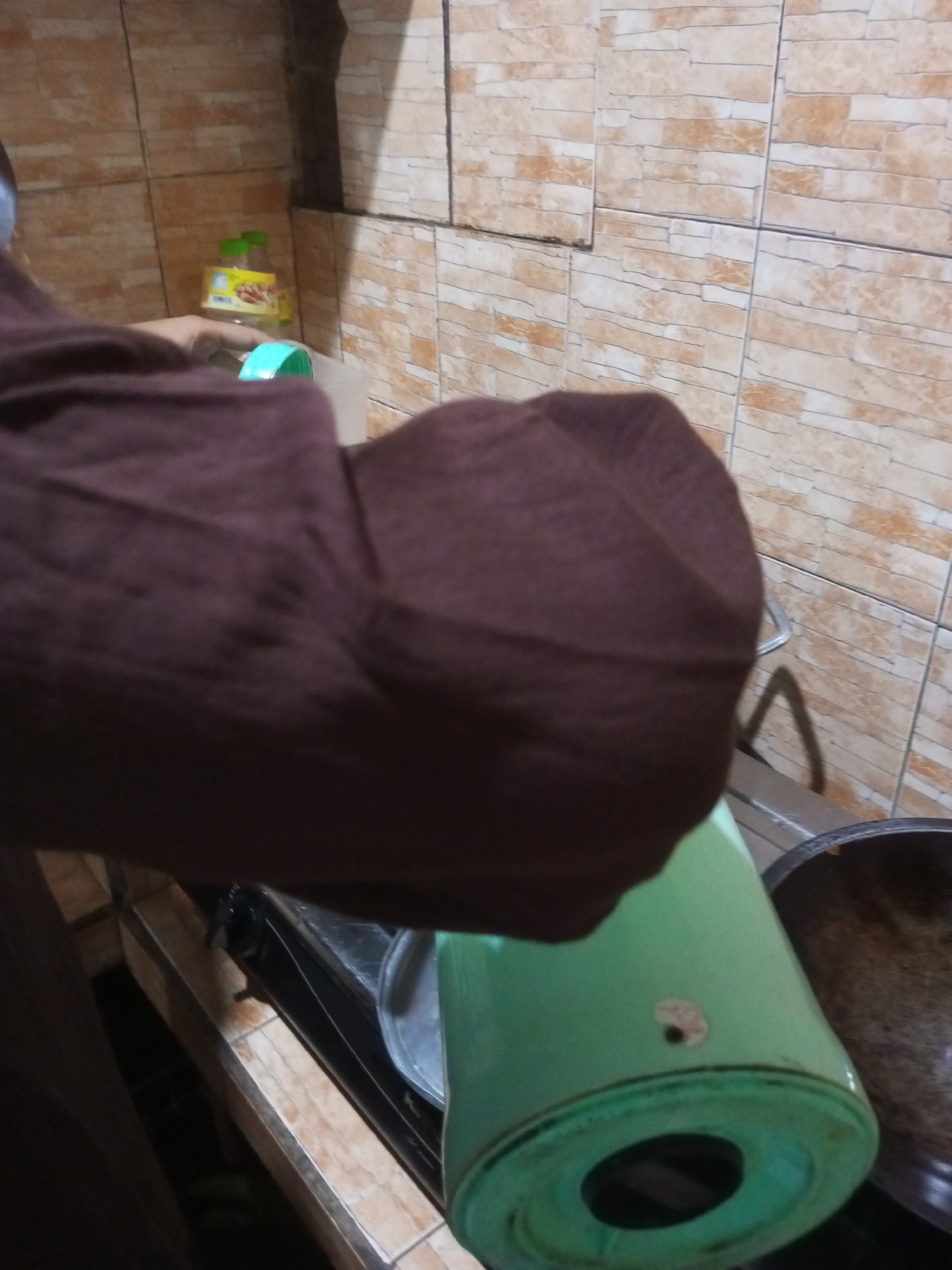 An overhead view captures a person's brown sleeve and part of their arm, positioned over a light green cylindrical object. The object, possibly a thermos or pitcher, has a hole in its base and a small sticker near the top. Behind the arm and object, a kitchen setting is visible. A tiled wall with a pattern of beige and orange rectangular tiles forms the backdrop. To the left, a small yellow bottle with a green cap sits on a shelf. Below, a black gas stove with a silver-rimmed pot is partially visible, indicating the scene is likely in a kitchen.
