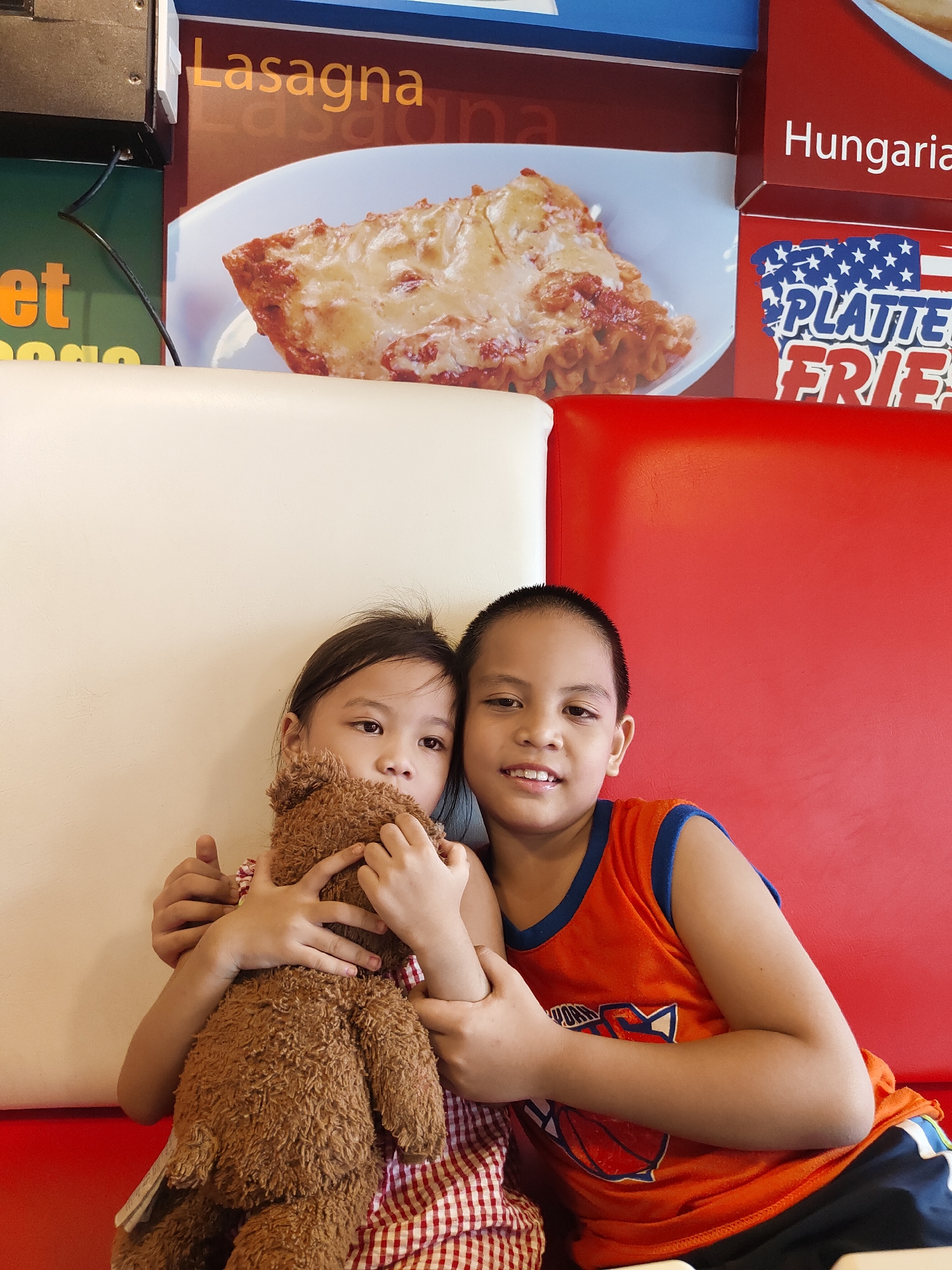 This is a medium shot photograph of two young children, a boy and a girl, seated next to each other on a red and white upholstered bench. The girl is on the left, holding a brown teddy bear tightly in her arms, with her face turned slightly away from the camera. The boy is on the right, smiling at the camera. Both children are looking towards the viewer. The background features promotional posters for food items, including "Lasagna," "Hungary," and "PLATTE Fries" with an American flag motif. The lighting appears to be artificial, suggesting an indoor setting, possibly a restaurant or fast-food establishment. The children's attire is casual: the girl wears a red and white checkered dress, and the boy is in an orange basketball-themed tank top and dark shorts. The overall mood is lighthearted and candid, with the boy's smile conveying happiness and the girl's embrace of the teddy bear suggesting comfort or affection. The image captures a moment of togetherness between the siblings.