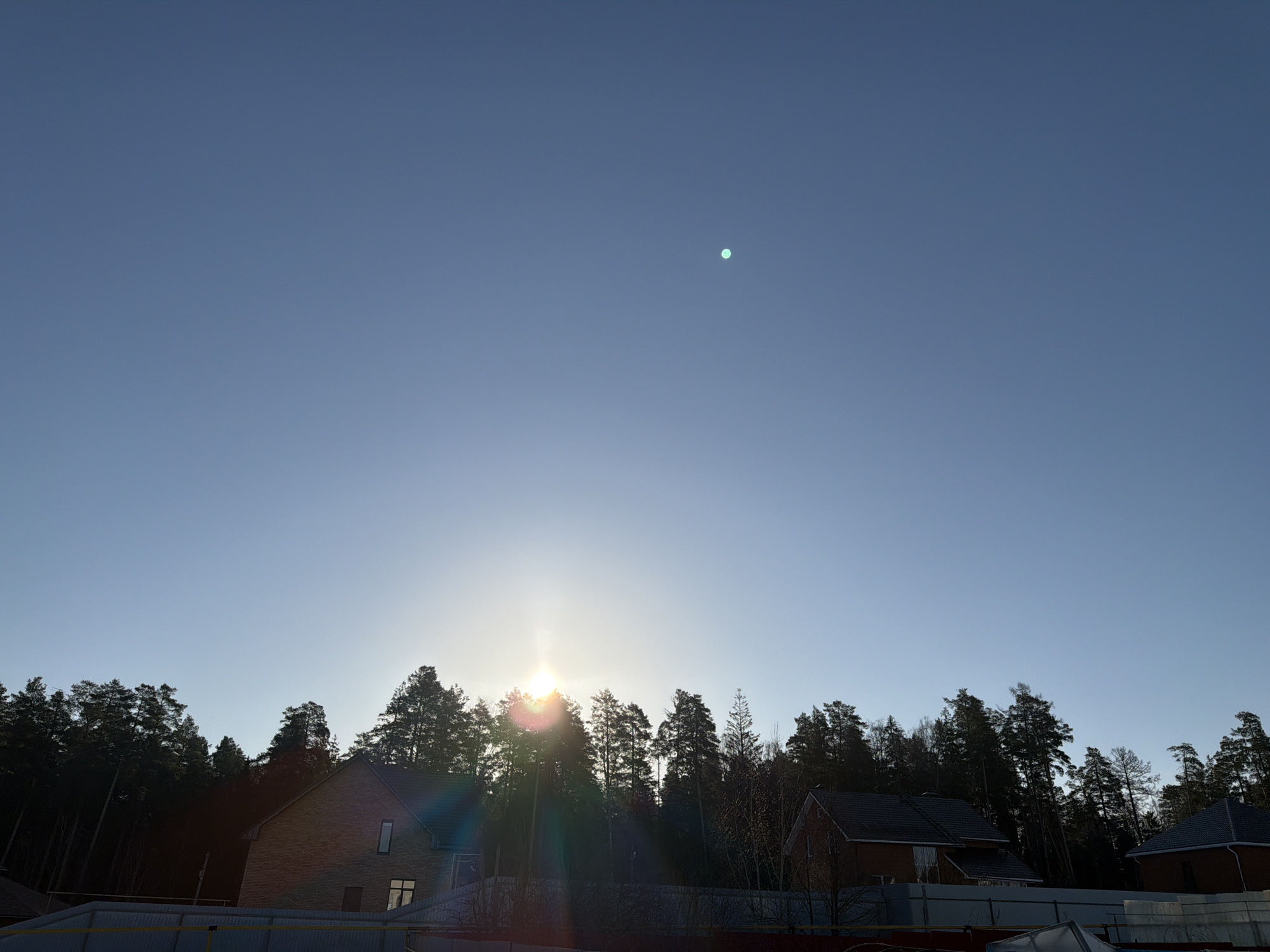 The image captures a wide-angle view of the sky above a forested area and a few houses. The location is Chaikovsky, Russia. The sky is a clear blue gradient, slightly lighter near the horizon where the sun is positioned, partially obscured by the treeline. A single bright spot is visible in the upper part of the sky. The treeline consists of tall pine trees, creating a dark silhouette against the bright sky. Below the trees, there are red brick houses with dark roofs, only partially visible. A fence runs along the bottom of the frame.