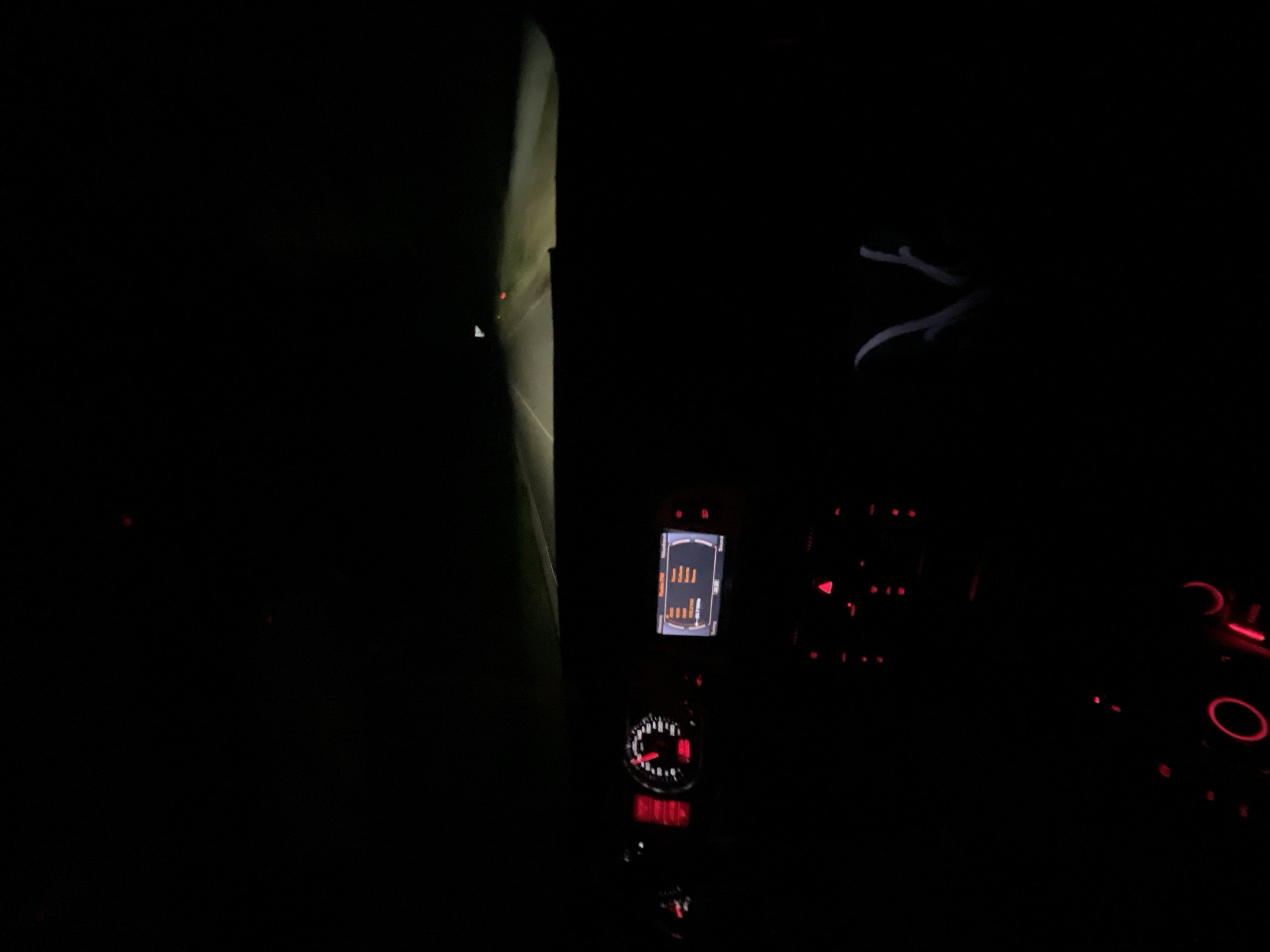 The image shows the interior of a car at night, with a view of the road ahead through the windshield. The dashboard is illuminated with red and white lights. A speedometer is visible on the left, showing a speed of approximately 90 km/h. In the center, a screen displays what appears to be radio information. Red buttons and controls are scattered across the dashboard. The road ahead is dimly lit by the car's headlights, with a few distant lights visible. The image is taken from the passenger seat. No specific location in Bogolin, Bulgaria can be determined from the image.