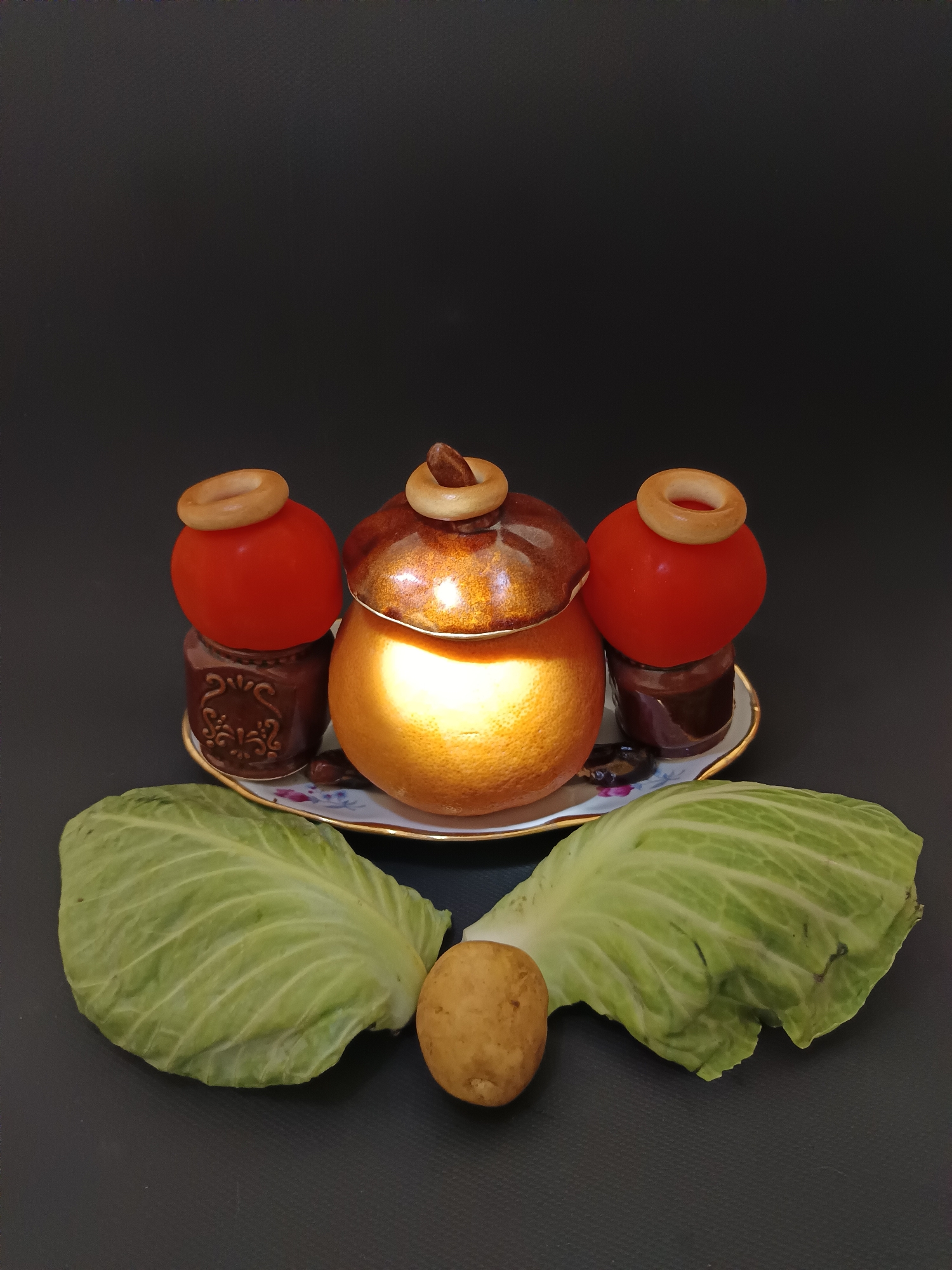 In Tyshky, Ukraine, a still life arrangement is set against a dark background. The scene features a decorative plate holding a large, orange-like object with a lid, illuminated by a strong light source. To the left and right of this central object are red, tomato-like spheres topped with round, light-brown rings. These spheres rest on smaller, dark brown, cylindrical objects. Partially visible on the plate are dark, bean-like items. In the foreground, two large, green cabbage leaves are spread out, with a single, small, light-brown potato positioned between them. The overall presentation is a staged still life.