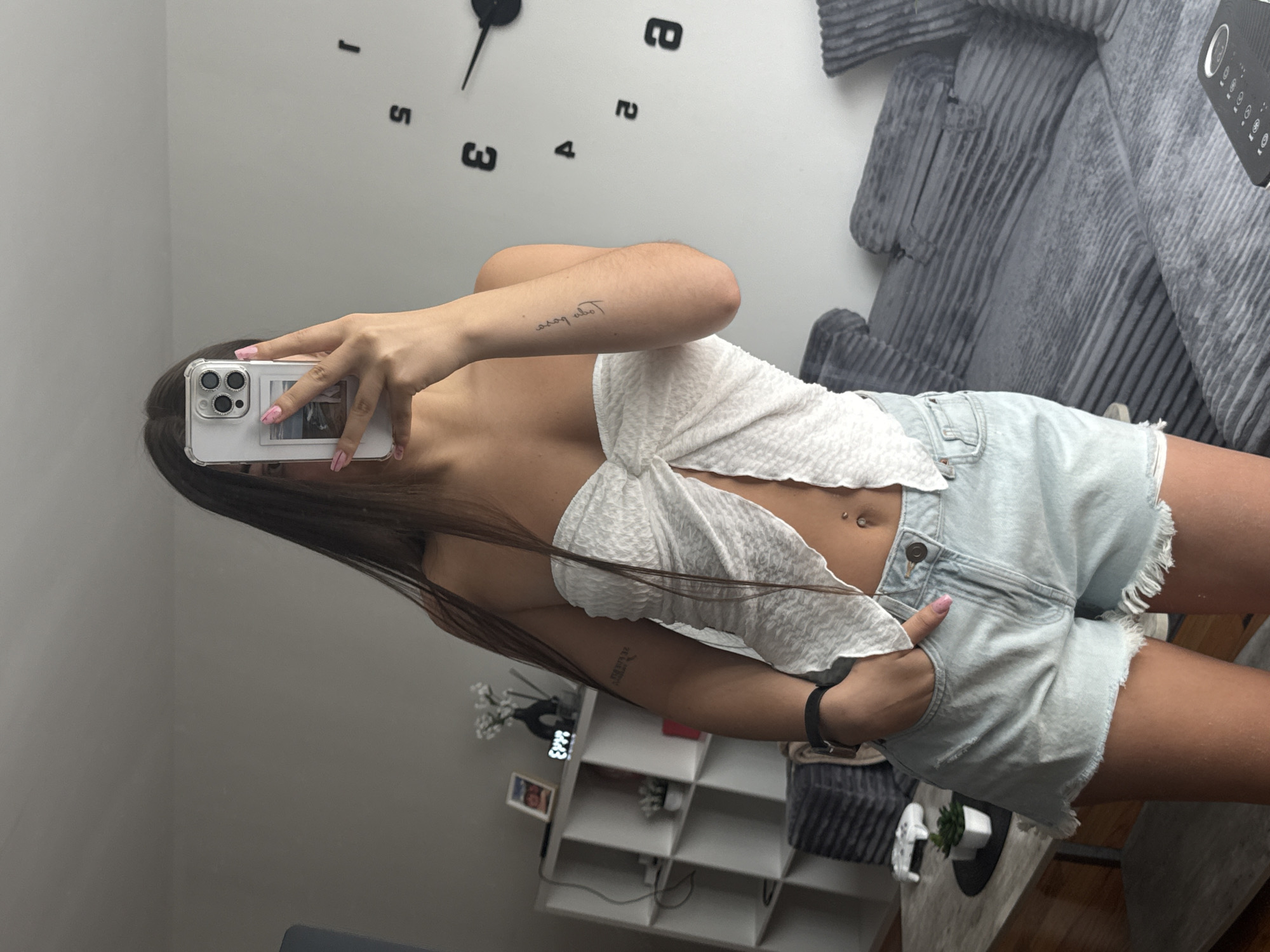 This media file is a vertical, full-body selfie taken in a bedroom. A young woman is the sole subject, holding up her smartphone to take the picture. She is wearing a white, textured, cropped top with a tie-front detail and light-wash denim shorts with a frayed hem. A navel piercing is visible. She has long, dark hair that falls over her shoulder. A small tattoo is visible on her left forearm.
The background reveals a modern bedroom setting. A large, graphic clock with black numerals is mounted on a light gray wall. To the right, a portion of a grey, ribbed sofa is visible. In the lower right of the frame, a white shelf unit with various items, including a vase with small white flowers, a framed picture, and a digital clock, is present. The overall lighting suggests it is daytime. No specific weather is indicated. There is no visible text beyond the numbers on the clock. The overall mood appears casual and relaxed.