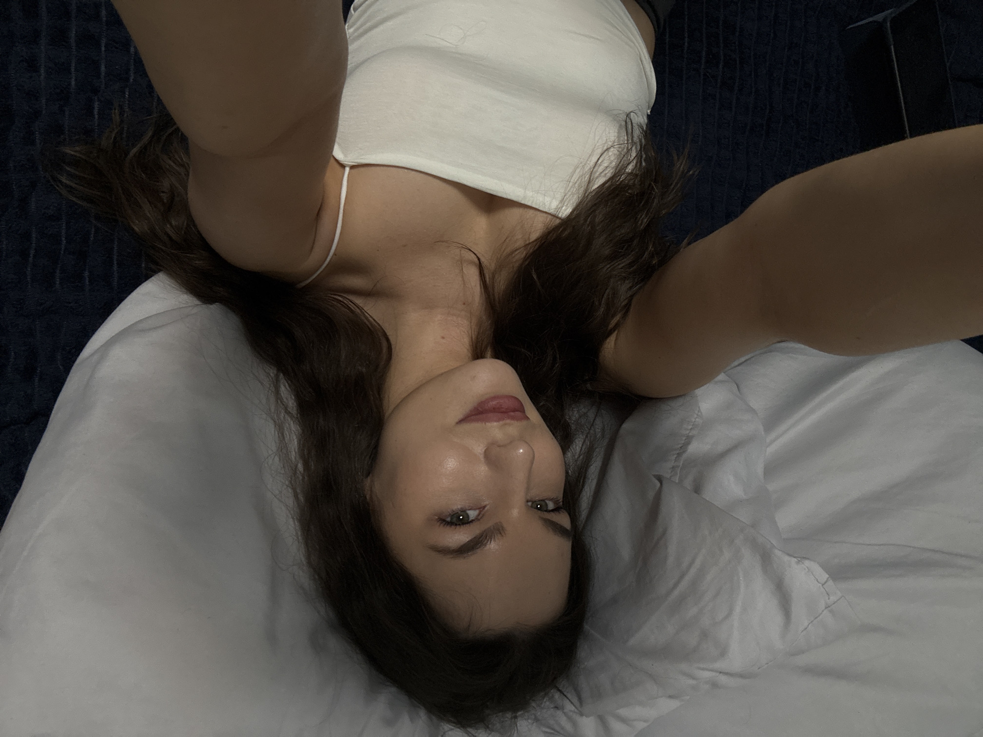 A young woman with long, dark wavy hair is lying in bed, looking at the camera. She is wearing a white spaghetti strap tank top. The background is a pillow and a dark blue, textured blanket. The scene appears to be indoors, likely a bedroom. There is no information to suggest this location is in Chaikovsky, Russia.