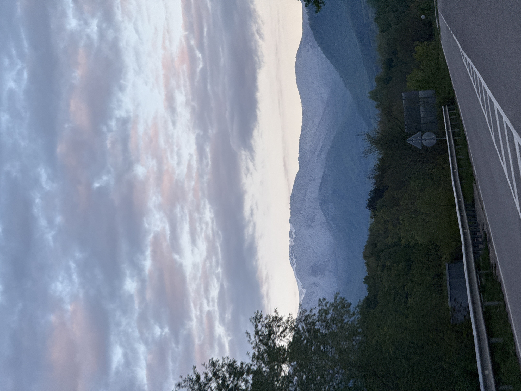 The image captures a serene landscape featuring a mountain range under a cloudy sky, with a portion of a highway in the foreground. The photograph appears to be taken during the golden hour, either at dawn or dusk, judging by the soft, diffused light and the delicate pink and orange hues visible within the cloud formations against a predominantly blue-gray sky.
In the mid-ground, a majestic mountain range dominates the horizon. The slopes of the closer mountains are covered in dense, dark green trees and foliage. Behind these, further peaks rise, appearing lighter in color, possibly indicative of snow cover or strong illumination from the low sun, and exhibiting a soft, muted blue-gray tone characteristic of atmospheric perspective.
The foreground on the right side of the image shows a section of an asphalt highway or road. It features distinct white lane markings, including a solid line along the edge and several dashed lines for traffic lanes. A metal guardrail runs parallel to the road. Beyond the guardrail and the road, a sloped area is densely covered with dark green trees and vegetation. Partially obscured within the foliage, a dark, rectangular structure, possibly a sign or a building, is faintly visible, along with a small, indistinct triangular road sign.
No discernible text is visible on any signs or objects. The overall scene conveys a sense of calm and natural beauty, characteristic of a mountainous region, in this case, Mezdreya, Bulgaria.