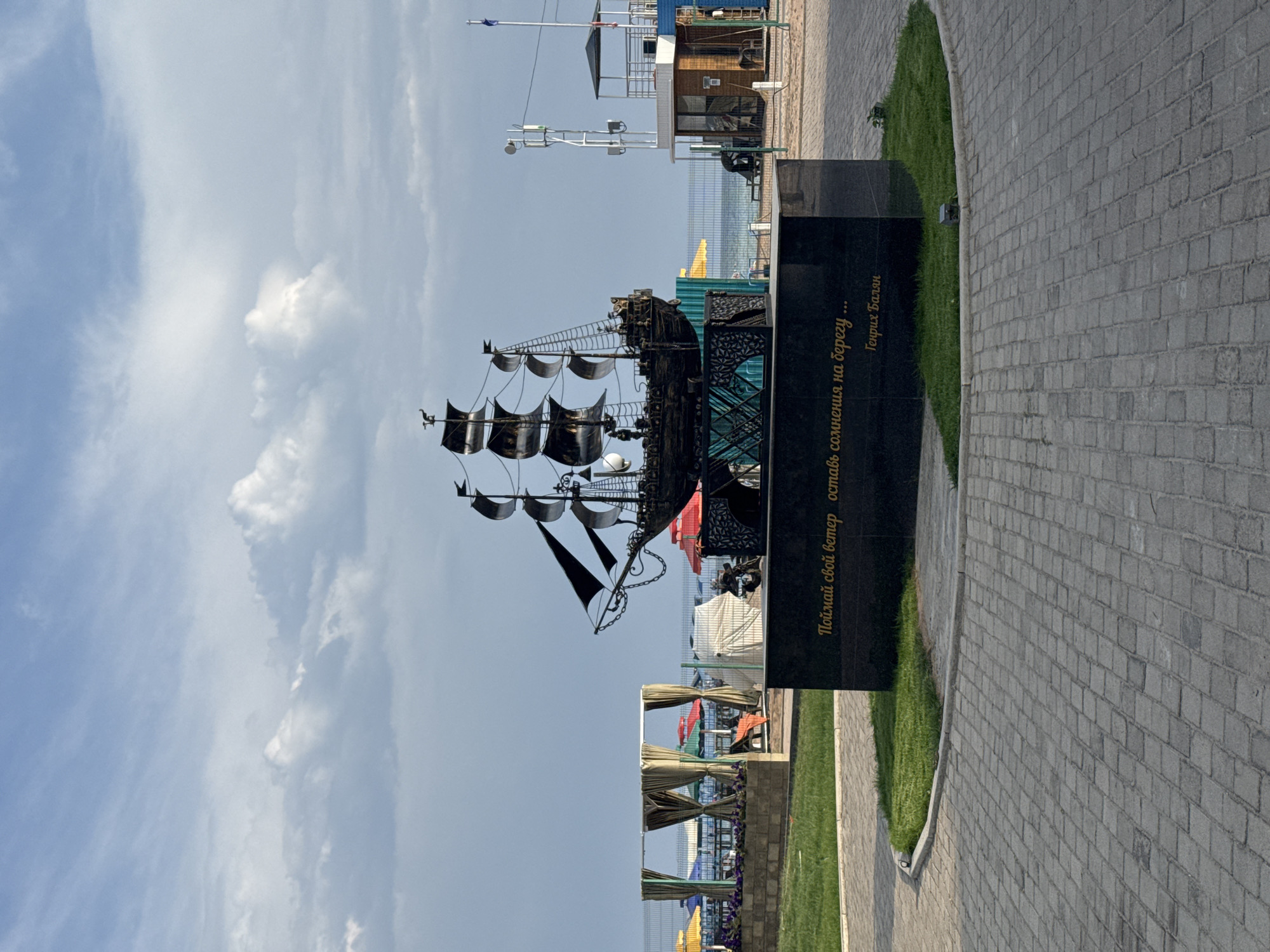 The image shows a metal sculpture of a sailing ship on a dark granite pedestal in a paved area. The sculpture is detailed, with masts, sails, rigging, and small figures on deck. The pedestal has golden lettering which reads "Поймай свой ветер оставь сомнения на берегу..." and "Генрих Бальян". In the background, beyond a metal fence, there is a beach with colorful umbrellas and lounge chairs. The sea is visible, and the sky is partly cloudy with blue patches. To the right of the sculpture, there is a lifeguard station or small building with a tower and antennae. The environment appears to be a recreational area, likely a resort or park, on the shore of a body of water. The location is identified as Sary-Oy, Kyrgyzstan.