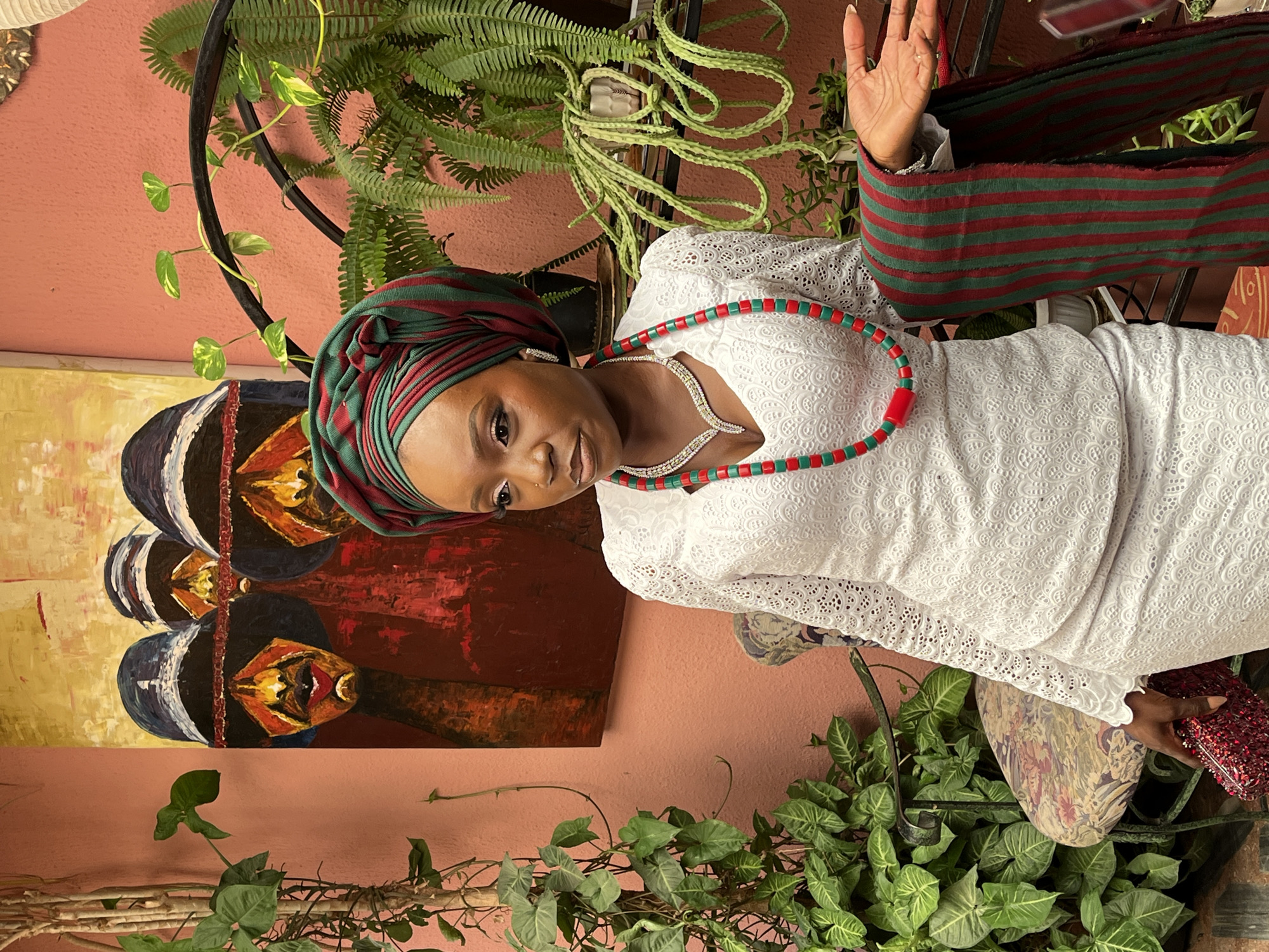 The image shows a young Nigerian woman dressed in traditional attire, standing in what appears to be a courtyard or an indoor garden. She is wearing a white lace outfit, a green and red headwrap, and matching beaded jewelry. A red, sequined clutch is in her left hand, and a striped scarf is draped over her right arm. Behind her, a painting with three figures and lush green plants are visible. The woman is looking off to the side with a neutral expression. The setting suggests a vibrant cultural context, possibly in Bar Jirgi Summa, Nigeria, given the traditional clothing and artistic elements.