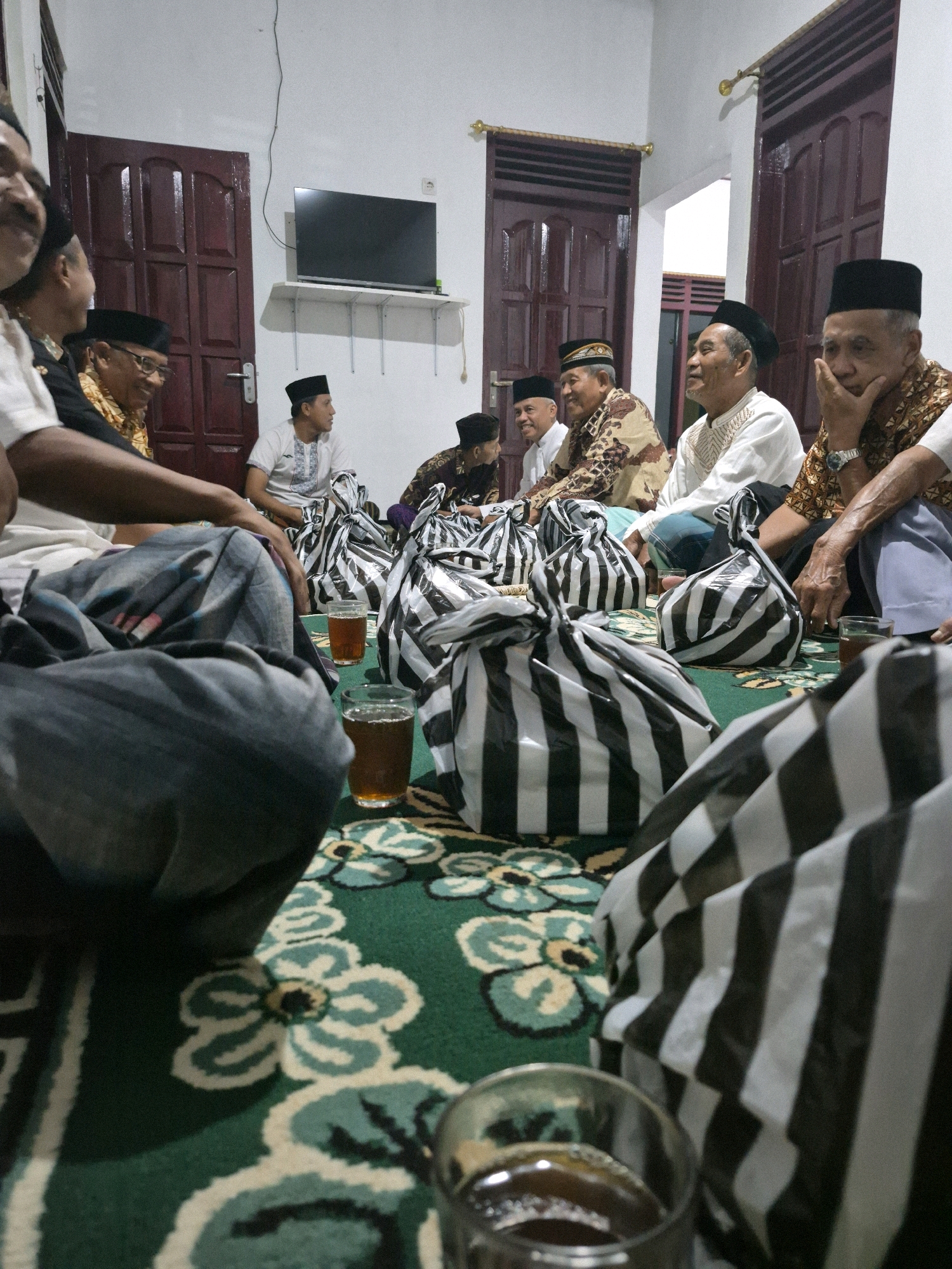 The image depicts a gathering of men, likely in a residential setting in Dukuhrejo, Indonesia, possibly for a religious or cultural event. They are seated on the floor, which is covered by a green carpet with a floral pattern. Several black and white striped bags are arranged on the carpet, some containing what appears to be food or gifts. Two glasses of a dark liquid, presumably tea or coffee, are visible on the carpet.
The men are dressed in traditional attire, including peci (caps) and patterned shirts or sarongs. Their expressions suggest a relaxed and communal atmosphere. The background shows a white wall with wooden doors and a television mounted on a shelf, indicating a domestic interior. The lighting is dim, suggesting the event is taking place indoors, possibly in the evening. The presence of multiple people and the communal seating arrangement suggest a social gathering, and the striped bags could be indicative of a celebration or distribution of items.