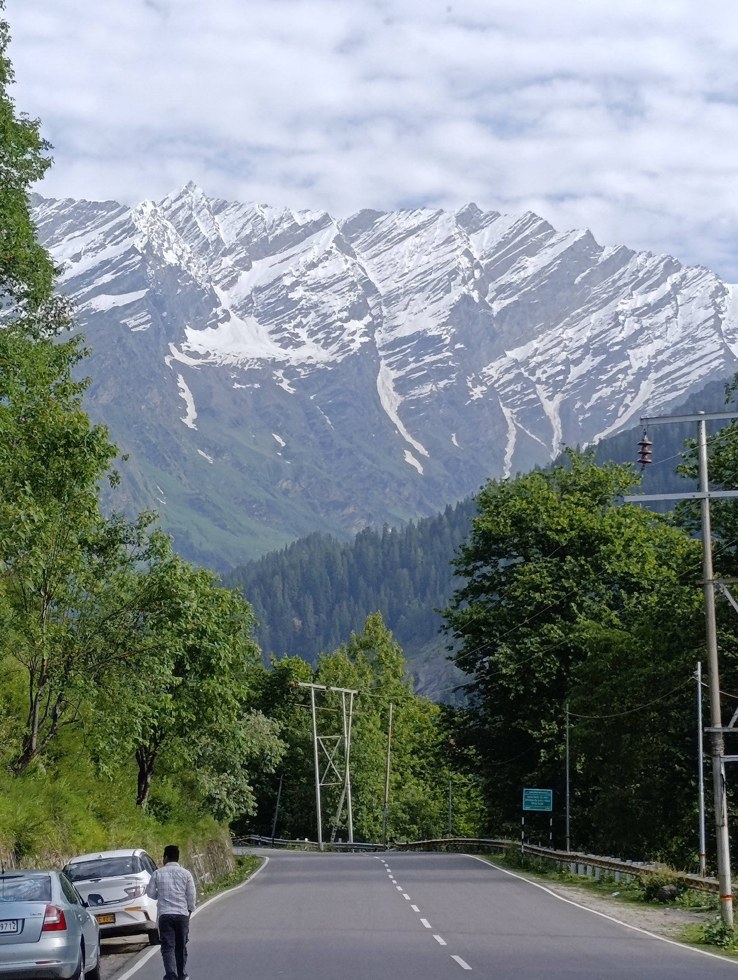 A full shot captures a paved, empty road winding through a green, mountainous landscape under a cloudy sky. Snow-capped mountains rise in the background, their rugged peaks outlined against the sky. The foreground shows a man walking away from the camera down the road, dressed in a checkered shirt and dark pants. Two cars are parked on the side of the road to the left, one silver and one white. Lush green trees line the sides of the road, with power lines and poles visible in the background. A green sign is posted on the right side of the road, and a guardrail runs along the edge of the road. The scene appears to be in Solang, India, known for its scenic beauty and adventure activities.