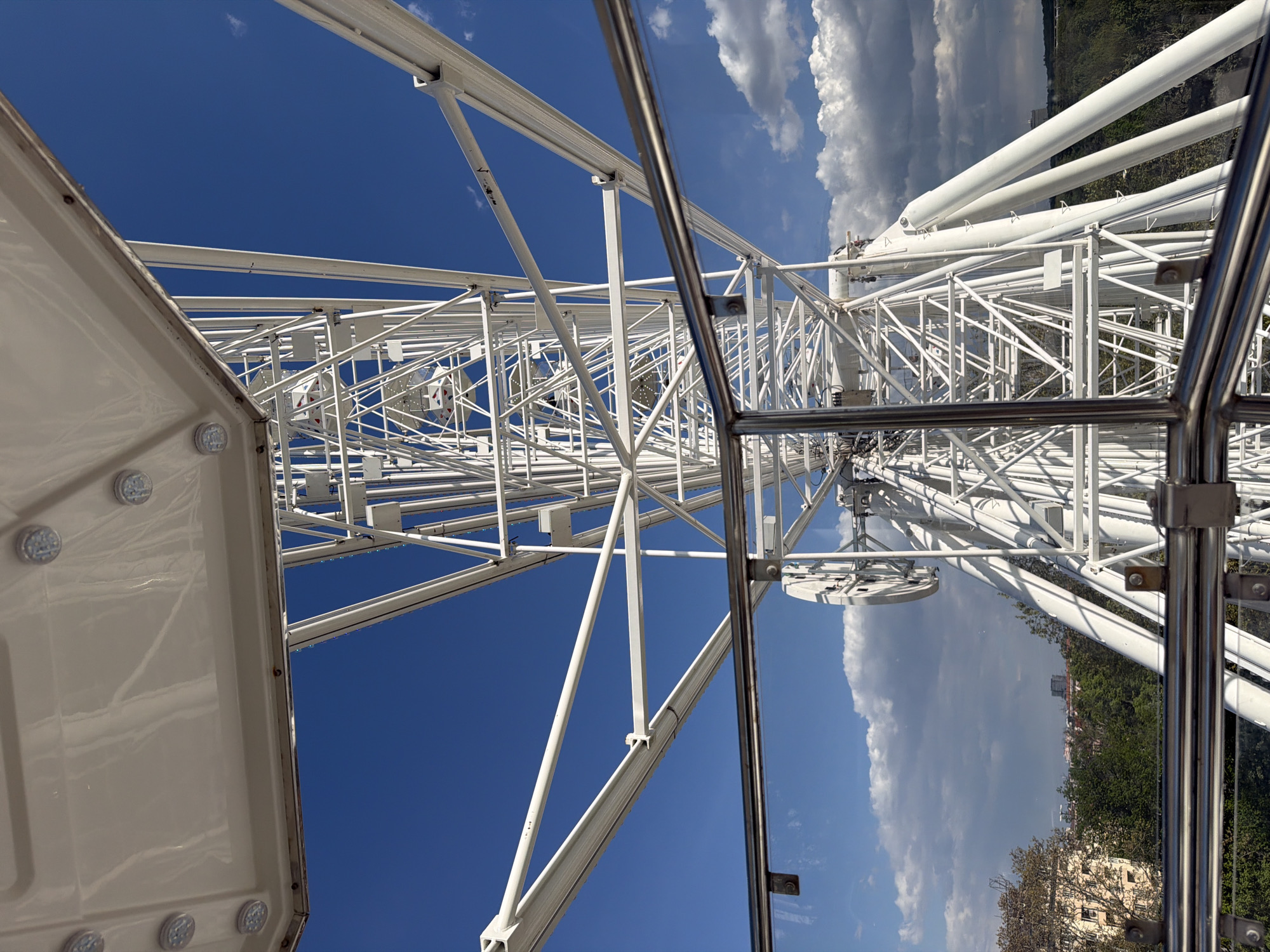 The image depicts a view from inside a Ferris wheel. The Ferris wheel's white metal framework, characterized by multiple angled supports and connecting rods, dominates the frame. A segment of the cabin is visible on the left, showcasing its white exterior and internal lights.
Outside the cabin, a vista of the sky and surroundings unfolds. The sky displays a vibrant contrast, featuring clear blue sections juxtaposed with fluffy white clouds. A glimpse of the city of Sofia, Bulgaria, is noticeable in the background, with treetops and building rooftops suggesting the urban environment.