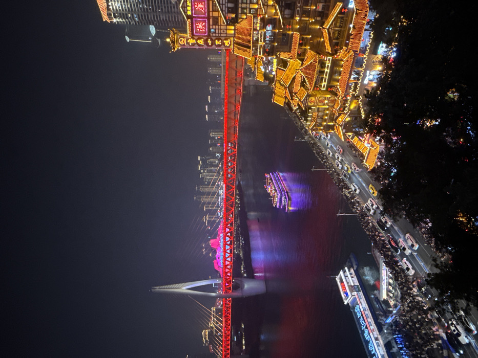 The image displays a nighttime urban scene in Yuzhong District, Chongqing, China, centered around the Jialing River, the Hongyadong complex, and the Qianximen Bridge. The entire area is extensively illuminated by artificial lights.
On the right side of the foreground, the Hongyadong complex, a multi-story traditional Chinese architectural structure, cascades down towards the river, brightly lit with warm yellow and orange lights. Chinese characters are visible on vertical signage elements. Directly in front of the complex, a road runs along the riverbank, populated by numerous vehicles, including cars and yellow taxis, many exhibiting illuminated headlights or taillights. A dense crowd of people occupies the promenade adjacent to the road, extending along the riverfront. Dark tree foliage with scattered colorful decorative lights is visible in the lower right.
The Jialing River dominates the midground, reflecting the diverse lights from the surrounding structures and vessels, creating areas of purple, blue, and red on its surface. A large, multi-decked cruise boat, brightly lit with purple and yellow illumination, navigates the center of the river. A second, smaller multi-decked boat, illuminated in blue and white, is positioned near the left riverbank, possibly docked, with individuals visible on its decks.
Spanning the river in the background is the Qianximen Bridge. Its main structure, a truss bridge, is vibrantly illuminated in red, as are its cables. A prominent white A-frame pylon supports the cable-stayed section of the bridge on the left. Beyond the bridge and across the river, a dense cityscape featuring numerous modern high-rise buildings is visible, illuminated against the dark night sky. The sky above is clear and dark, indicating nighttime conditions.
