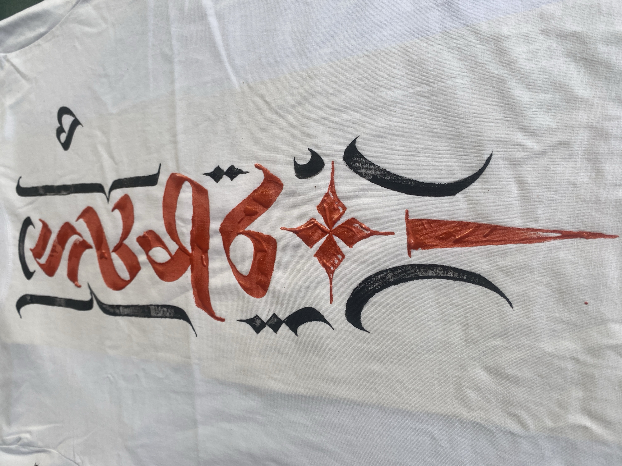 On a white cloth, likely a shirt, a decorative design is painted in black and orange. The design appears to be inspired by Tibetan or similar calligraphic styles, featuring stylized characters and symbols. A small heart-like shape is visible at the top left. The cloth is wrinkled. The photograph was taken at Baan Nai Suan, Thailand.
