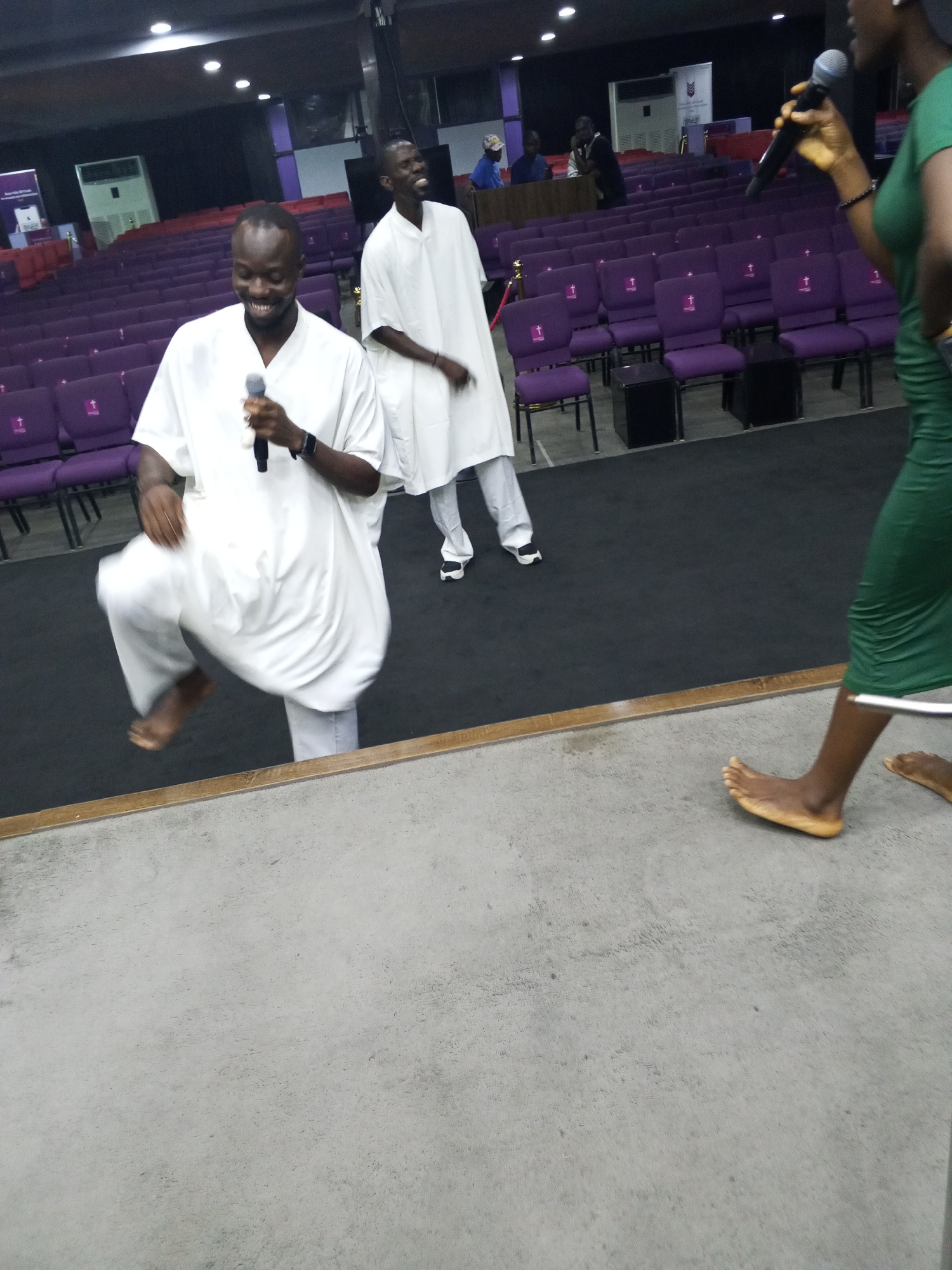 The image shows three individuals on a stage in what appears to be an auditorium or lecture hall. The location is unknown. The hall has rows of purple chairs with a white cross symbol on the back.
A man in the foreground is wearing a white, flowing garment. He is holding a microphone and appears to be in motion, possibly dancing. Another man in a similar white garment is visible behind him, also holding a microphone. A woman is partially visible on the right side of the frame. She is wearing a green dress and is barefoot, also holding a microphone.
The stage floor is gray and appears to be made of concrete. There are other people seated in the hall in the background, near a podium.