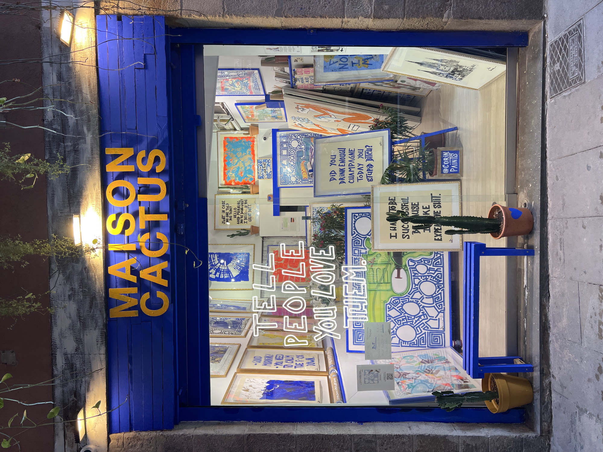 This full-shot image displays the brightly lit storefront of an art gallery named "MAISON CACTUS" at night, located in Barcelona, Spain, confirmed by the "SOCIEDAD GENERAL DE BARCELONA" inscription on a manhole cover in the foreground.
The facade features a dark, weathered upper section from which green vines hang, illuminated by two rectangular white LED lights. Below, a vibrant royal blue wooden paneling serves as the backdrop for the shop's name, "MAISON CACTUS," rendered in large, bold, golden-yellow uppercase letters. The shop's entrance is a large glass window framed in the same royal blue.
Inside the well-lit store, numerous artworks are displayed, predominantly featuring blue, white, and occasional orange or yellow tones, often with bold graphic patterns or text. A prominent white neon sign in the center of the window reads, "TELL PEOPLE YOU LOVE THEM." Other visible text-based artworks include: "GOOD MORNING! YOU NEED TO CALM THE F*CK DOWN." (partially visible on the left), "JUST IN CASE NO ONE TOLD YOU TODAY HELLO GOOD MORNING YOU ARE DOING GREAT I BELIEVE IN YOU GREAT BUTT." (top left), "DID YOU DRINK ENOUGH CHAMPAGNE TODAY YOU STUPID BITCH?" (center-right), and "I HAVE TO BE SUCCESSFUL BECAUSE I LIKE EXPENSIVE SHIT." (partially visible bottom right). A small blue sign near the floor in the bottom right indicates "@CORIN PAINTS." Several potted cacti are placed throughout the interior display.
On the grey paved sidewalk directly in front of the shop, a royal blue wooden bench is positioned centrally. To the left of the bench, a tall cactus in a mustard yellow pot sits on the ground. To the right, another tall cactus in a terracotta pot, adorned with a small blue heart, stands. No individuals are present in the scene.