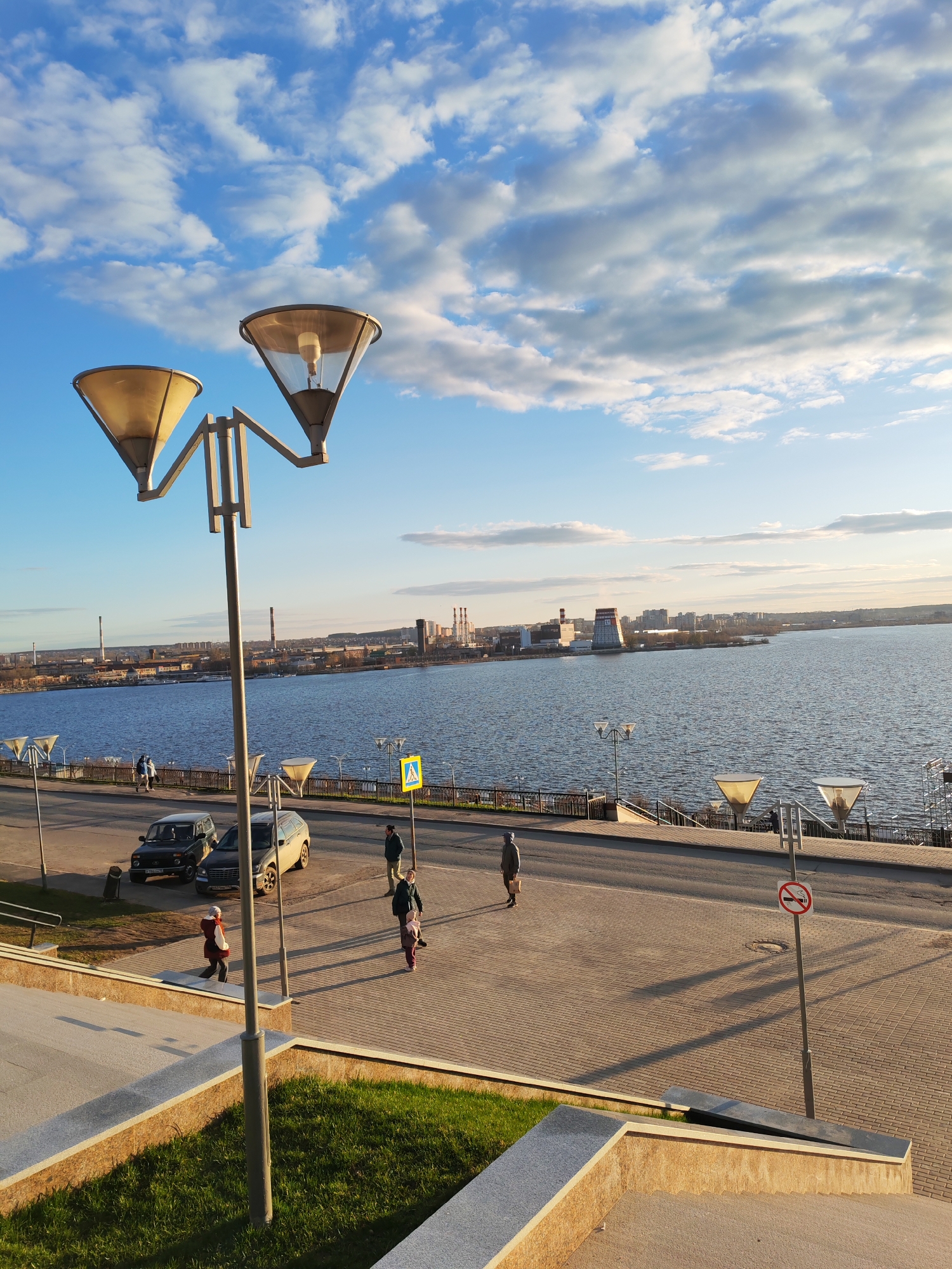The image depicts a waterfront scene in Shaberdino, Russia, on a sunny day with scattered clouds in a blue sky. The viewpoint is elevated, looking down onto a paved promenade bordering a wide river. In the middle distance, across the river, is an industrial landscape with smokestacks and buildings.
Several individuals are present on the promenade. A woman with two children is walking on the left. Further along, a group of three people are walking, and a lone individual walks near a "no smoking" sign on the right.
Two cars, a dark SUV and a lighter-colored station wagon, are parked along the roadside. Street lamps with distinctive inverted conical shades are spaced along the promenade. In the foreground, a grass patch is bordered by concrete steps. A pedestrian crossing sign is visible in the distance.