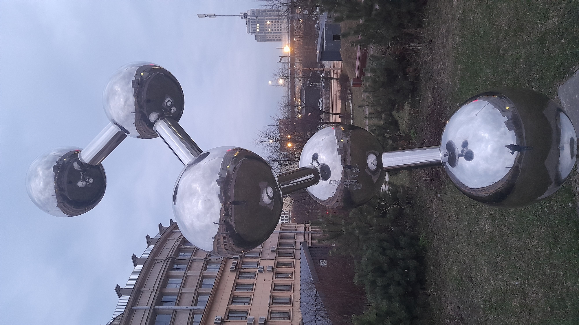 The image shows a large, shiny, metallic sculpture of a molecule in an outdoor setting. The sculpture consists of four interconnected spheres supported by cylindrical arms. The reflective surfaces of the spheres mirror the surrounding environment, including buildings and the sky.
In the background, there is a multi-story building with numerous windows, suggesting it could be an apartment complex or an office building. Beyond the sculpture, there is a street with streetlights, and in the distance, a taller building is visible. The ground around the sculpture is covered with grass and some bushes or small trees.
Based on the architectural style and cityscape, the image is likely taken in Saint Petersburg, Russia.