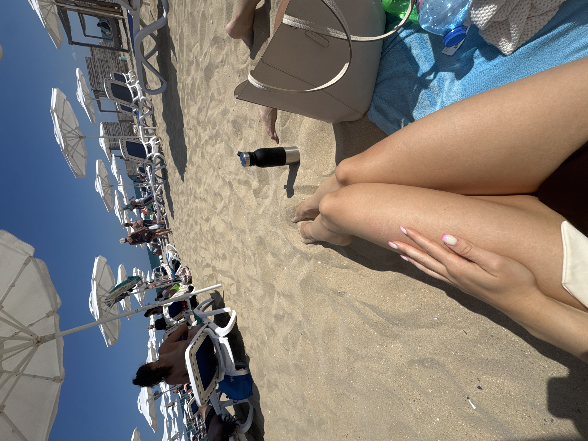 The image depicts a sunny day at a beach resort in Nesebar, Bulgaria. The foreground is dominated by the legs and hand of a person reclining on a blue towel, with a light-colored beach bag and a black water bottle nearby. In the mid-ground, rows of white beach umbrellas and loungers are set up on the sand, with several people visible relaxing and walking. The background shows a clear blue sky and a glimpse of the Black Sea. The overall scene suggests a typical vacation setting at a seaside resort.