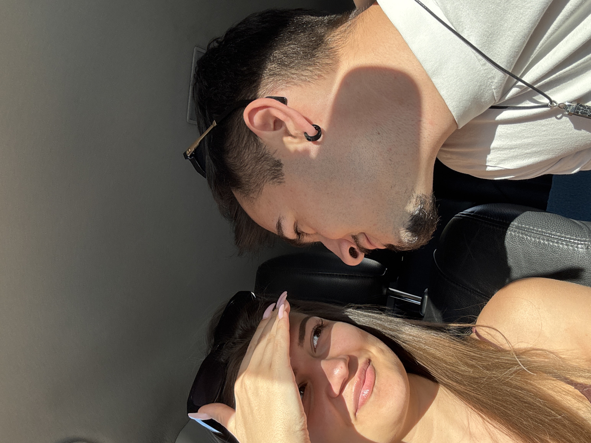 A photograph depicts two individuals inside a vehicle, potentially in transit. A woman with long brown hair and manicured nails is holding her hand up to shield her face from the sun, smiling slightly. She is wearing sunglasses pushed atop her head and a light-colored top. The man next to her has dark hair, a goatee, and an earring. He is wearing a white t-shirt and a necklace. The interior of the car includes black leather seats and a light-colored headliner. The location is not discernible as Sofia, Bulgaria without additional context.