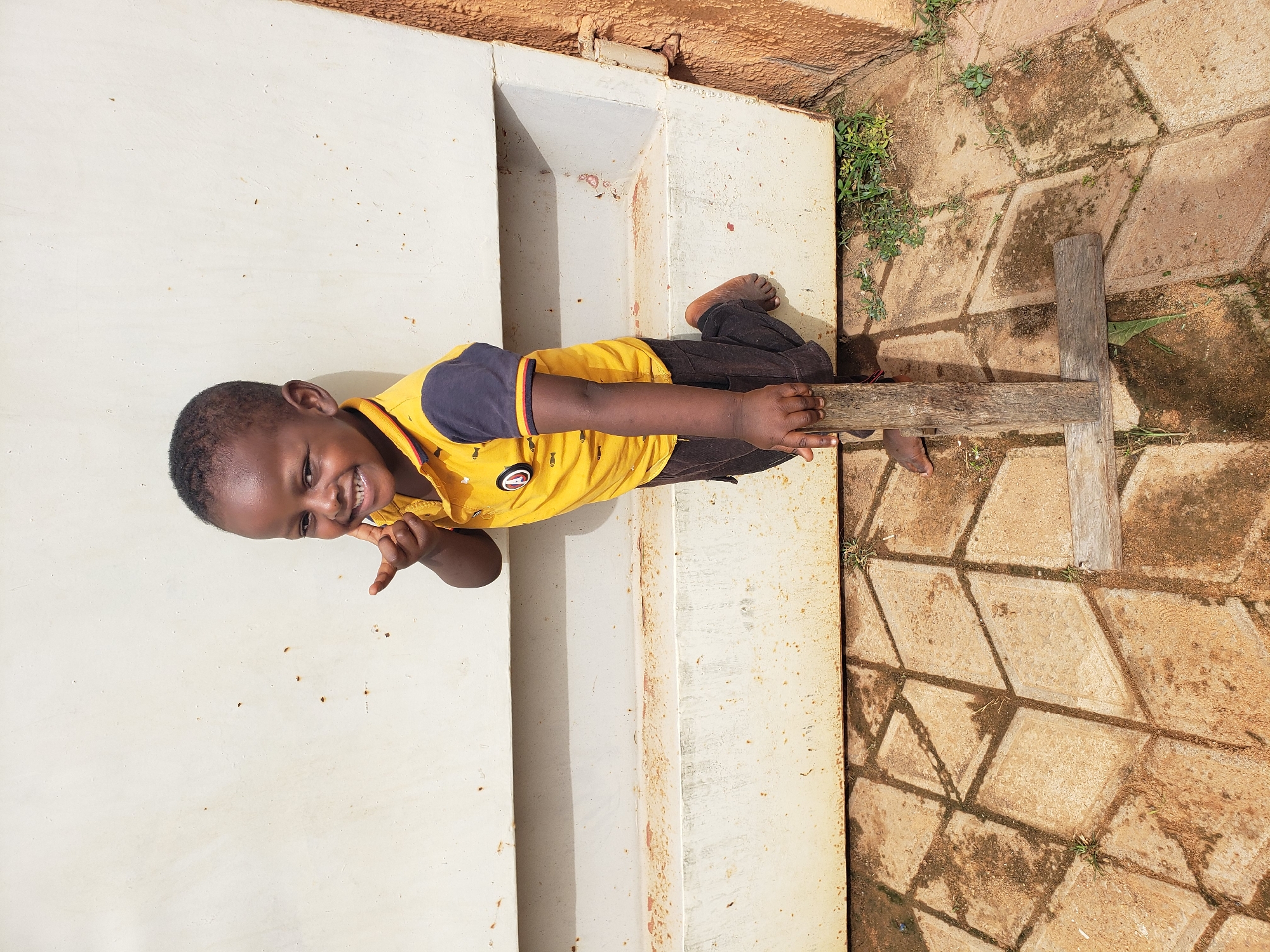A young Black child, appearing to be about 3-4 years old, stands with their right leg bent at the knee, propped up on a wooden object. The child is wearing a yellow t-shirt with navy blue sleeves and collar, featuring a small black and red circular logo on the left chest. The child has short black hair and is smiling directly at the camera, with their right index finger touching their cheek. The child is positioned in front of a large, off-white metal dumpster or wall, which shows signs of rust and dirt. The ground is paved with irregularly shaped, tan concrete tiles, some of which are cracked and overgrown with small green weeds. Shadows from the child and the dumpster fall across the tiled surface. The overall environment suggests an outdoor urban setting, likely in Sabon Tasha, Nigeria, given the context. The wooden object the child is propping their leg on appears to be made of two pieces of roughly cut wood, forming an 'L' shape.