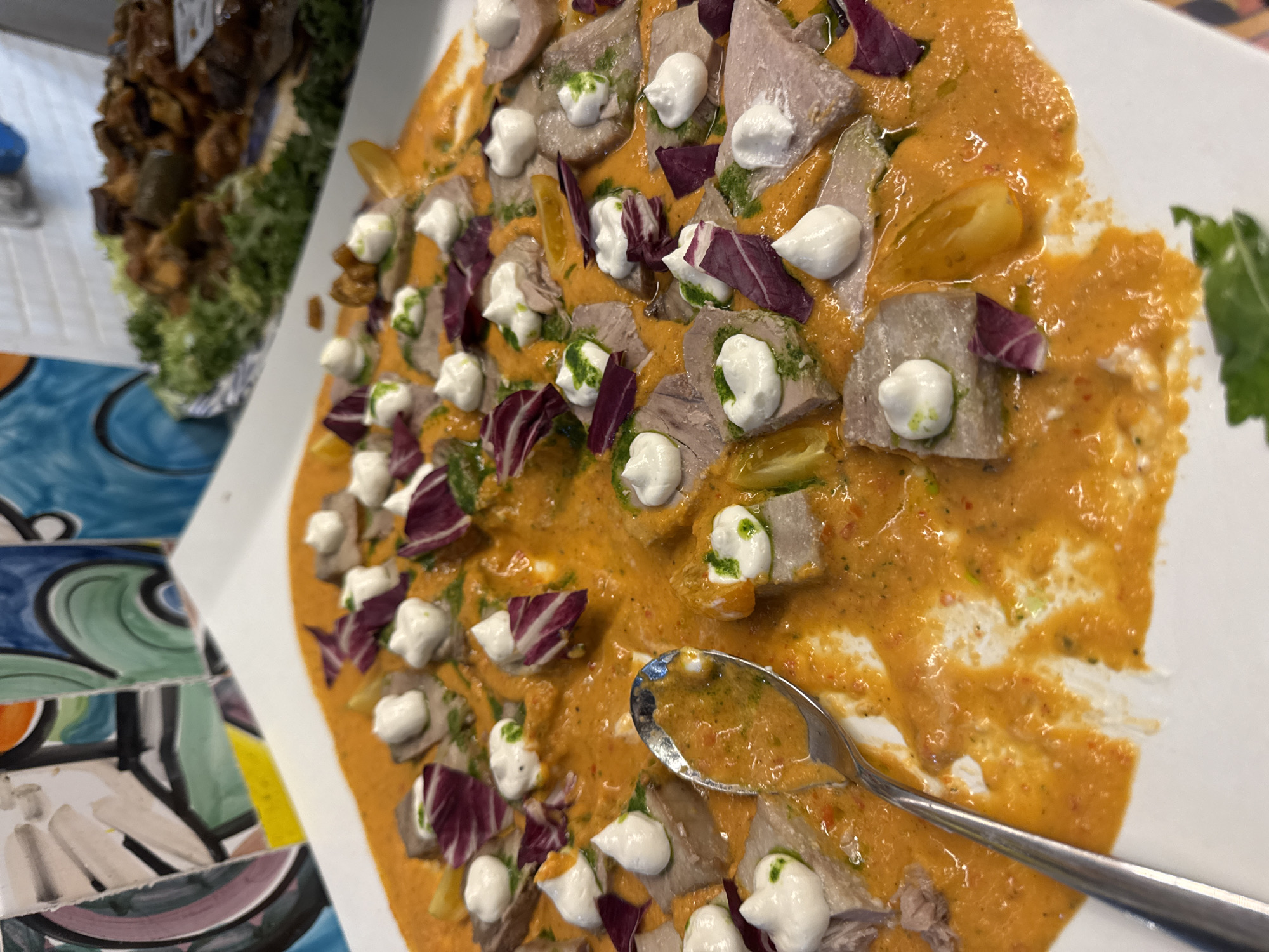 The image shows a close-up of a dish of food served on a white platter. The dish appears to be a carpaccio or thinly sliced meat covered in a vibrant orange sauce. Small dollops of white cream, possibly ricotta or a similar cheese, are placed on top of some of the meat slices, along with small pieces of red radicchio and a drizzle of green sauce, likely pesto or herb oil. A silver spoon is visible in the lower left corner, resting on the platter and partially submerged in the sauce. In the background, there is a blurred view of colorful tiles and another platter of food, which appears to be a vegetable dish. The overall presentation suggests a restaurant or catering setting, likely in Isola delle Femmine, Italy, given the context of the prompt. The lighting is bright, highlighting the colors and textures of the food.