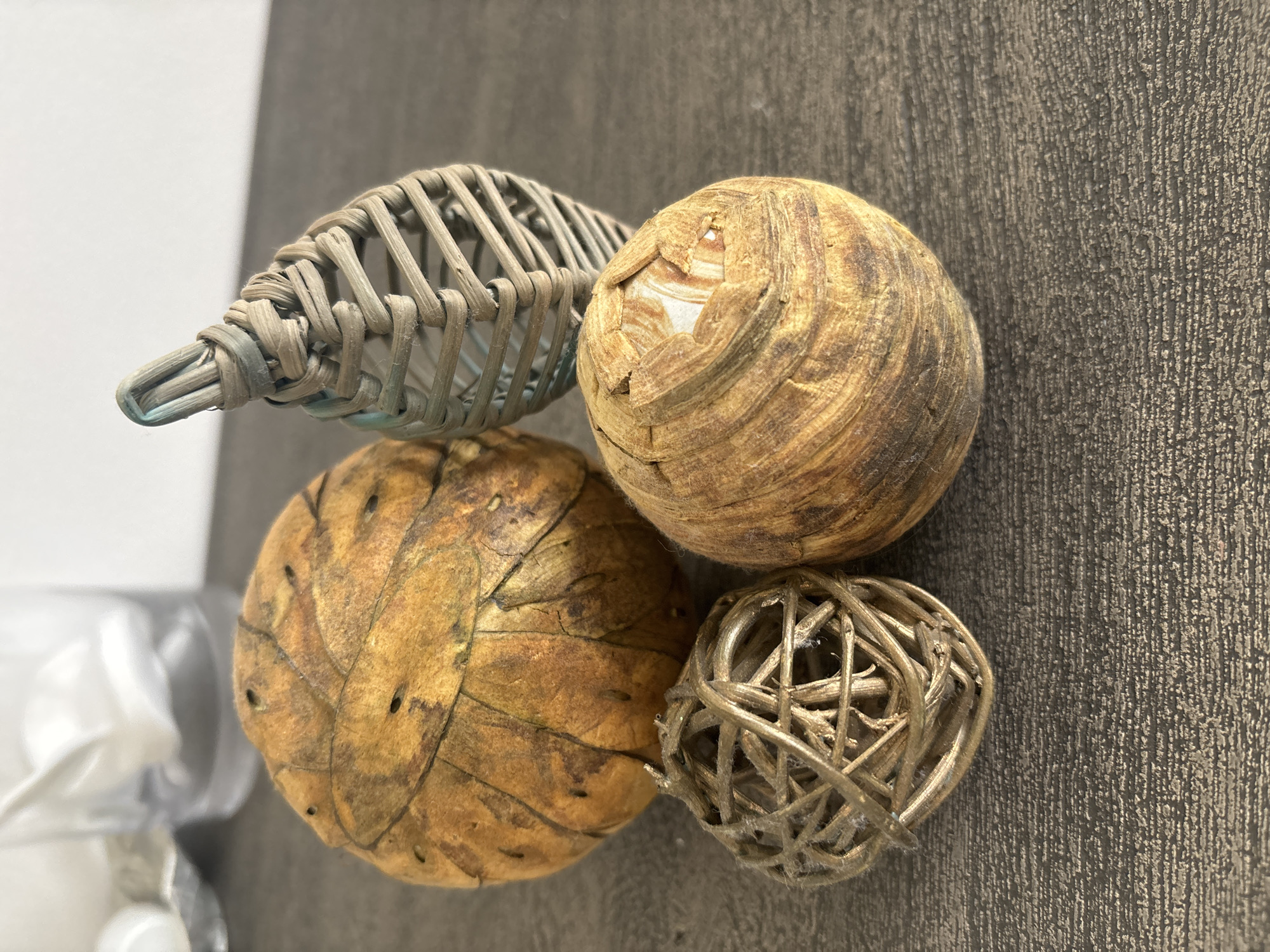 The image displays a close-up view of four decorative orbs and a woven, cone-shaped object resting on a dark, textured surface that appears to be a tabletop. The orbs vary in size and material. One large orb, in the upper left, is made of a dried, papery material with visible veins and small perforations. To its right, a smaller orb, in the foreground left, is constructed from thin, golden-brown twigs or fibers intricately woven into a spherical shape. A third orb, in the foreground right, is also spherical and appears to be made from a layered, fibrous material with a rough, textured surface, exhibiting variations in color from light beige to dark brown. A fourth object, to the right of the largest orb, is a tall, cone-shaped item made of tightly woven light-colored fibers, possibly rattan or straw, with a slightly bluish tint at its apex. The background is blurred, showing what appear to be clear glass containers and a bright white surface. The overall lighting suggests an indoor setting with natural light. There are no people or animals present in the image. The scene is static and appears to be a still life arrangement. The location in the city of Dubai, United Arab Emirates, cannot be determined from the visual information provided in the image.