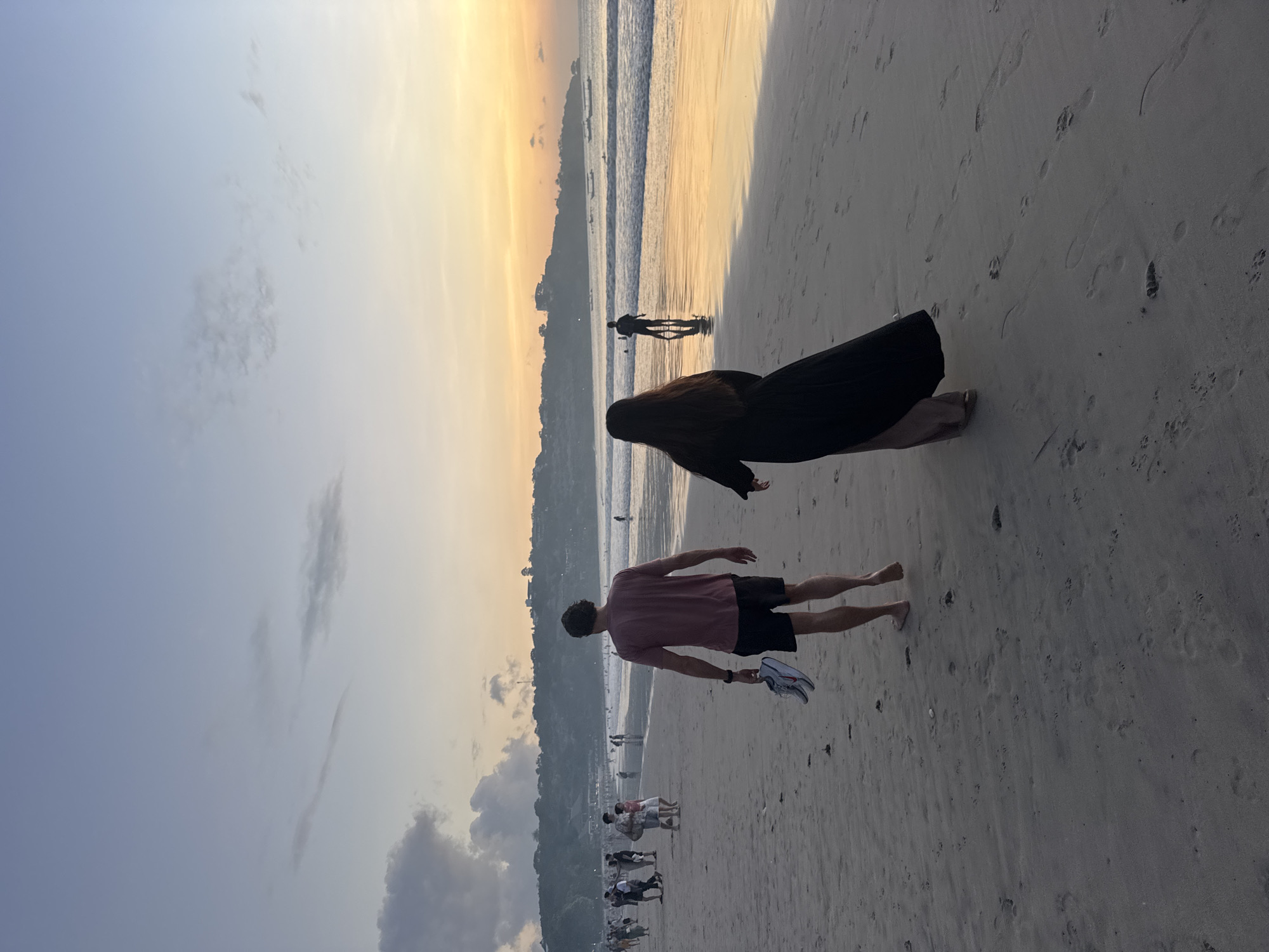 The image shows a beach scene at sunset in Kuta Selatan, Indonesia. A man in a pink shirt and black shorts is walking barefoot on the sand, carrying shoes in his left hand. Behind him, a person in a black garment is kneeling on the sand. Further back, near the water, is another person standing in the surf. There are several other people on the beach in the background near a tree covered land mass and an ocean in the background. The sky is a gradient of blue and orange from bottom left to top right.