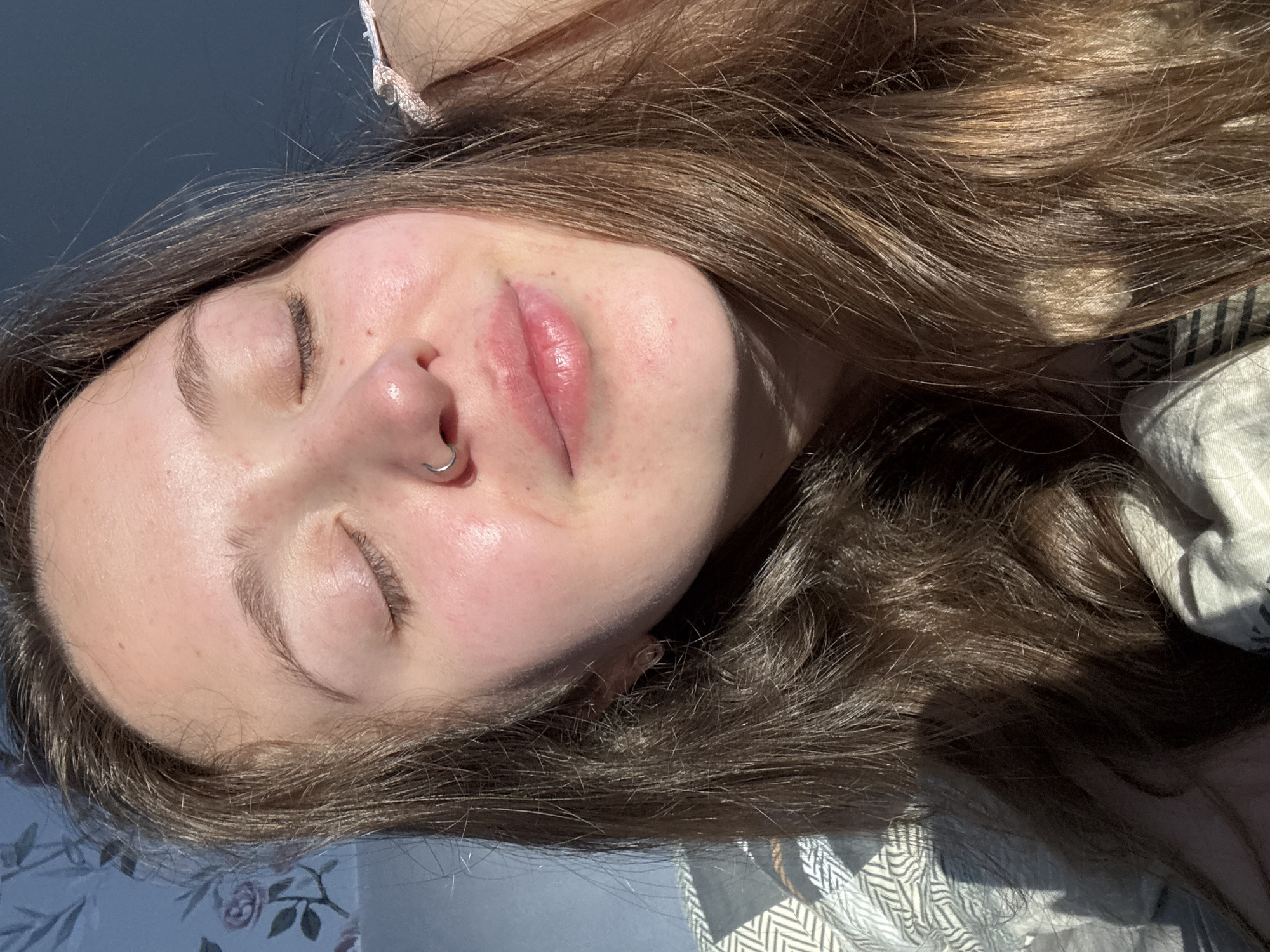 A person with closed eyes and light brown hair is lying down. They have a nose ring in their left nostril. The background includes a blue surface with a floral pattern and a white patterned fabric. The scene is brightly lit. There is no information to verify the location.