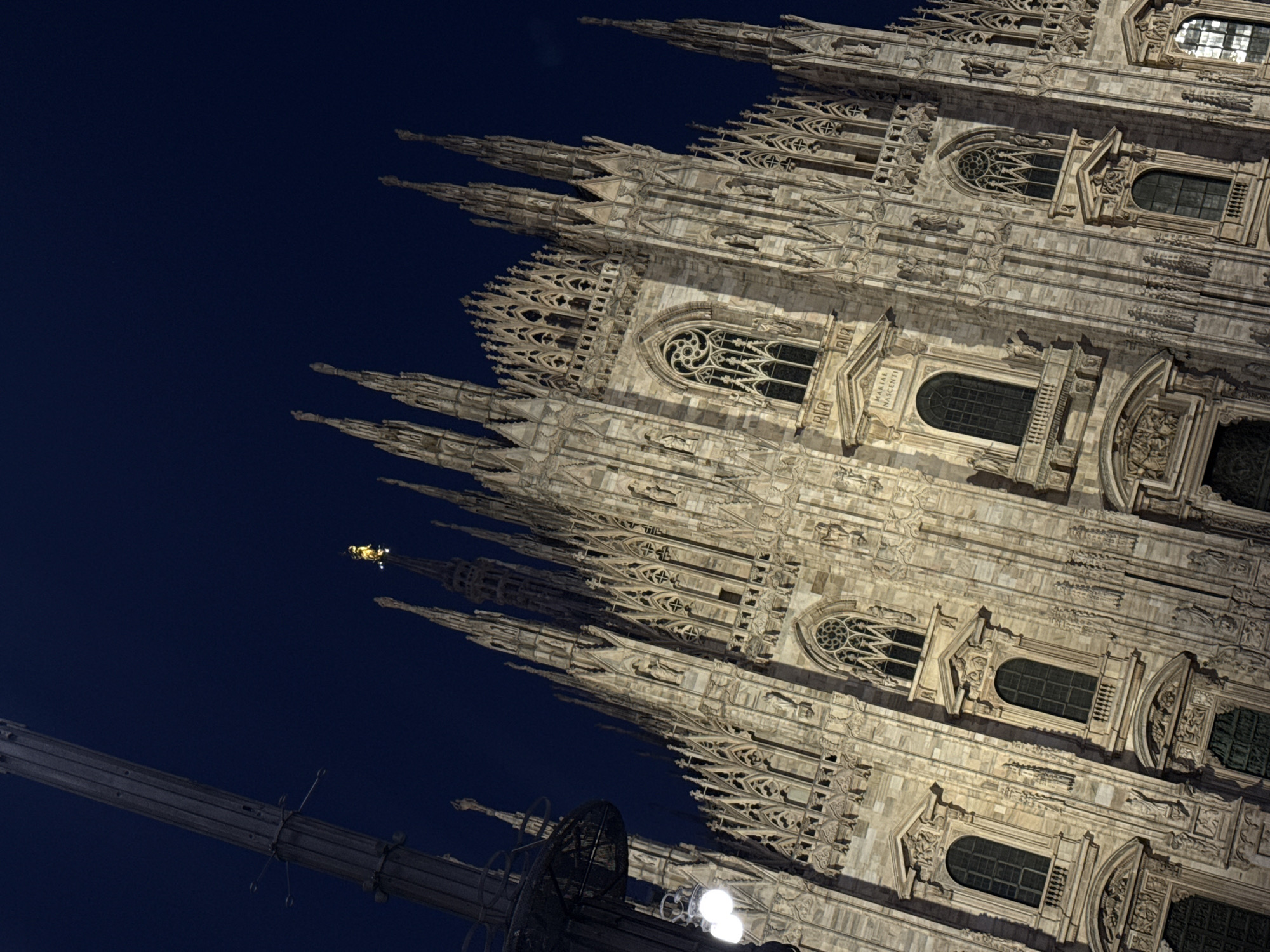 The image shows the facade of the Milan Cathedral (Duomo di Milano) at night. The building is intricately decorated with spires, statues, and Gothic-style windows. The sky is a dark blue, and the cathedral is illuminated. A metal pole-like structure is visible in the lower-left corner. The perspective is from a low angle, looking up at the structure.