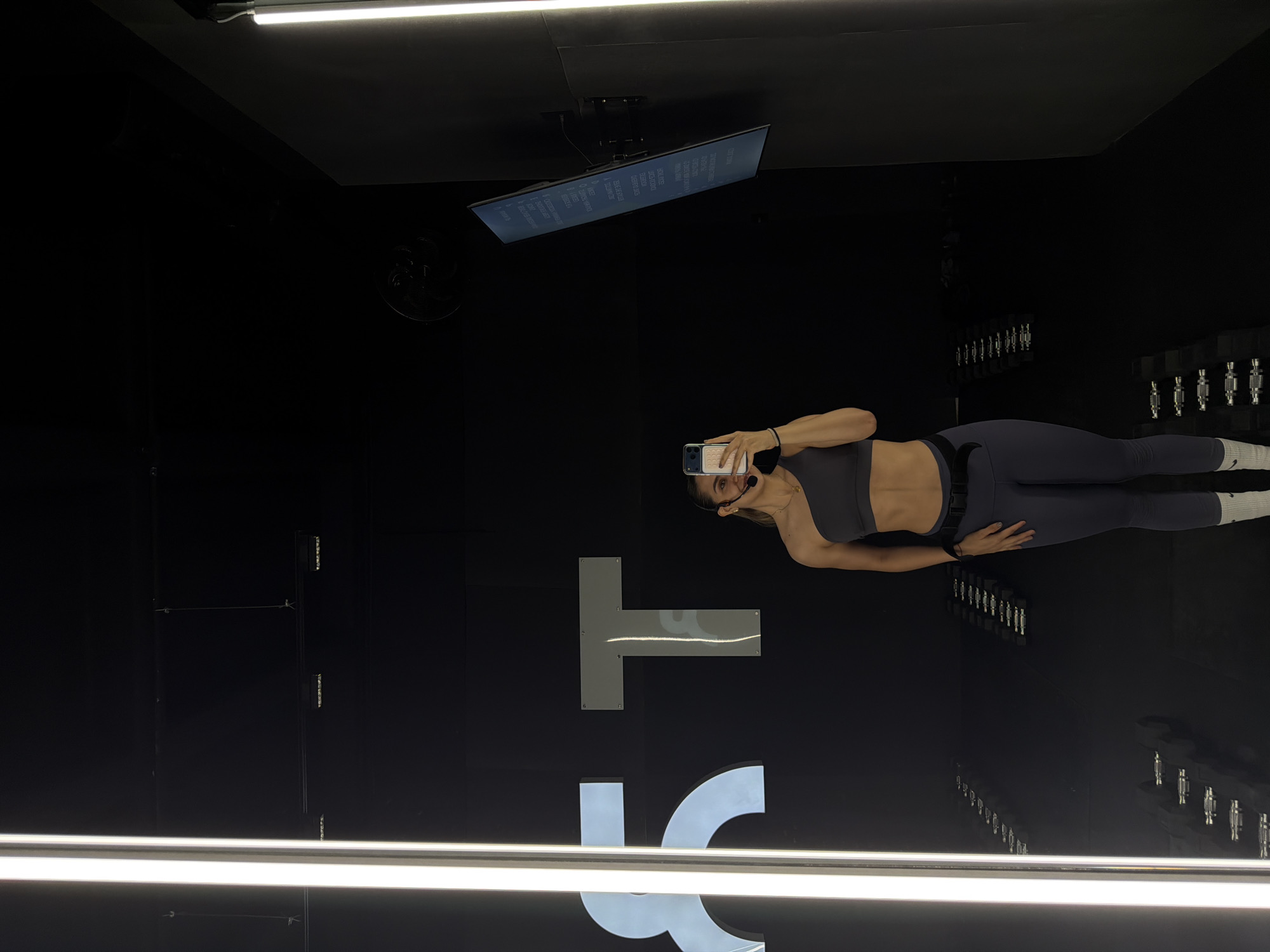 A woman is standing in front of a black wall with large white lettering. She is wearing a gray sports bra and leggings. She is holding a mobile phone and taking a picture of herself. A TV screen is mounted on the ceiling. Rows of weights are affixed to the wall. The location is Puebla, Mexico.