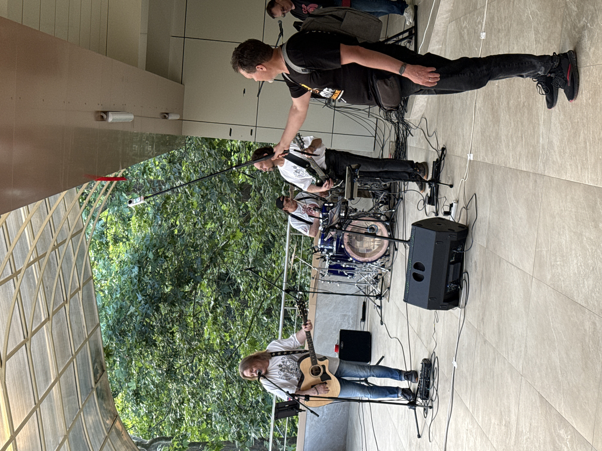 An outdoor concert is taking place under a covered walkway, with lush green trees visible behind the performers. A band of three men is on stage. The lead singer, a fair-skinned man with long blond hair, wears a white t-shirt and jeans and plays an acoustic guitar. To his right, another man in a white t-shirt and black pants plays an electric guitar. Behind them, a third man in a black t-shirt and jeans plays the drums. In the foreground, a fourth man, dressed in a black t-shirt and jeans, holds a boom microphone on a pole, extended towards the band. He has a black crossbody bag slung over his shoulder and wears a smartwatch. The stage is set up on a paved area with tiled flooring. Various audio equipment, including speakers and cables, are visible on the ground. To the far right, a fifth person, partially visible, stands with a backpack.