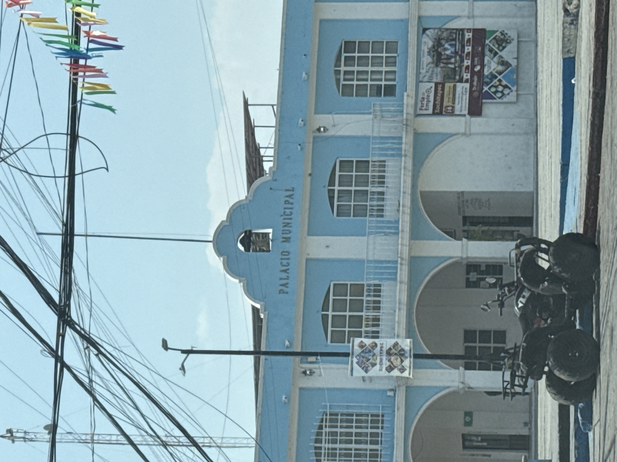 The image captures the Palacio Municipal (Municipal Palace) of Xochitepec, Mexico, on a clear, bright day. The building, painted in light blue with white trim and architectural accents, is a multi-story structure with a series of arched openings on the ground floor forming a portico. The upper floors feature a mix of arched and rectangular windows with white grilles. A prominent decorative element at the top center of the facade houses a bell, and the words "PALACIO MUNICIPAL" are displayed in white letters below it.
In front of the building, on the right side, several black ATVs (All-Terrain Vehicles) are parked. The sky is bright blue with faint clouds. On the left side of the image, numerous black electrical wires and cables crisscross overhead. A tall pole adorned with strings of colorful small flags or pennants is visible on the far left, suggesting a festive or celebratory atmosphere.
Two large banners are affixed to the building's facade on the upper right. One banner advertises a "Feria de Empleo" (Job Fair) and clearly states "Xochitepec" and "Trabajo" (Work), with the number "16" also visible. A smaller, square banner featuring similar imagery and text for "Xochitepec" hangs from a street lamp pole in the lower foreground. The scene depicts a public building bustling with potential activity, possibly related to the advertised job fair or a local event.