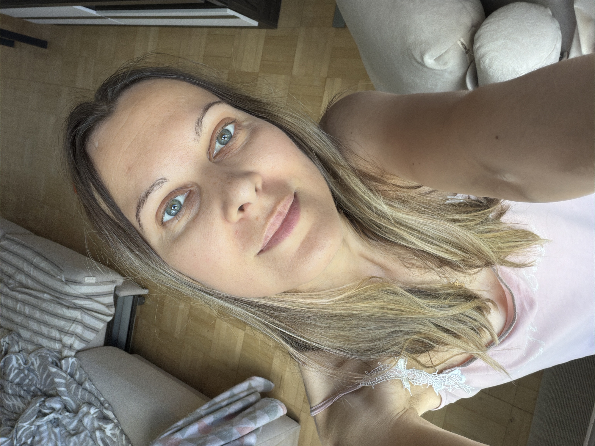 A selfie is taken from a high angle, looking down at a woman with blonde hair and blue eyes. She is wearing a pink camisole with white lace detailing on the straps. Her hair is long and wavy, falling over her shoulders. The background shows a room with wooden flooring and furniture. There is a couch with striped and patterned cushions, and a large, light-colored object that appears to be a stuffed animal or pillow. The lighting suggests it is daytime. There is no discernible information about the location being Izhevsk, Russia.
