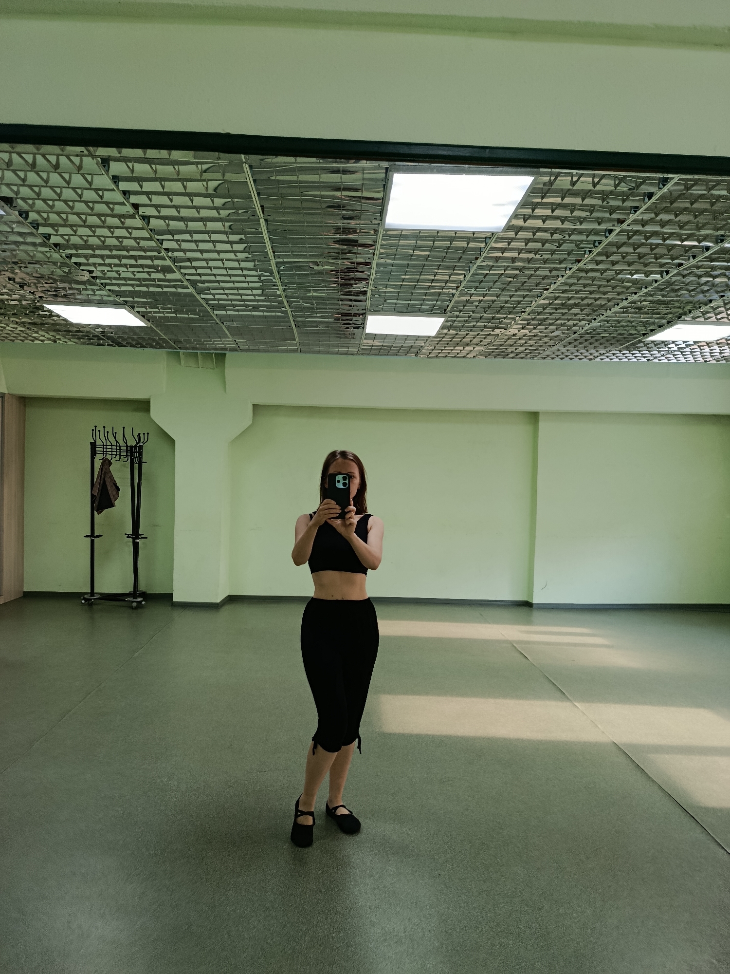 A young woman stands in a dance studio, taking a mirror selfie. She is wearing a black crop top and black capri pants, with black flat shoes. Her hair is long and brown, and she is holding a smartphone in her hands, partially obscuring her face. The studio has a light green painted wall and a grey floor. The ceiling is a grid of metal with recessed fluorescent lighting panels. A coat rack with several hangers and a brown garment is visible in the background to the left. Sunlight streams in through unseen windows, casting long shadows across the floor. The scene appears to be taking place during the daytime, as indicated by the bright lighting and shadows. The overall impression is one of a practice or rehearsal space.