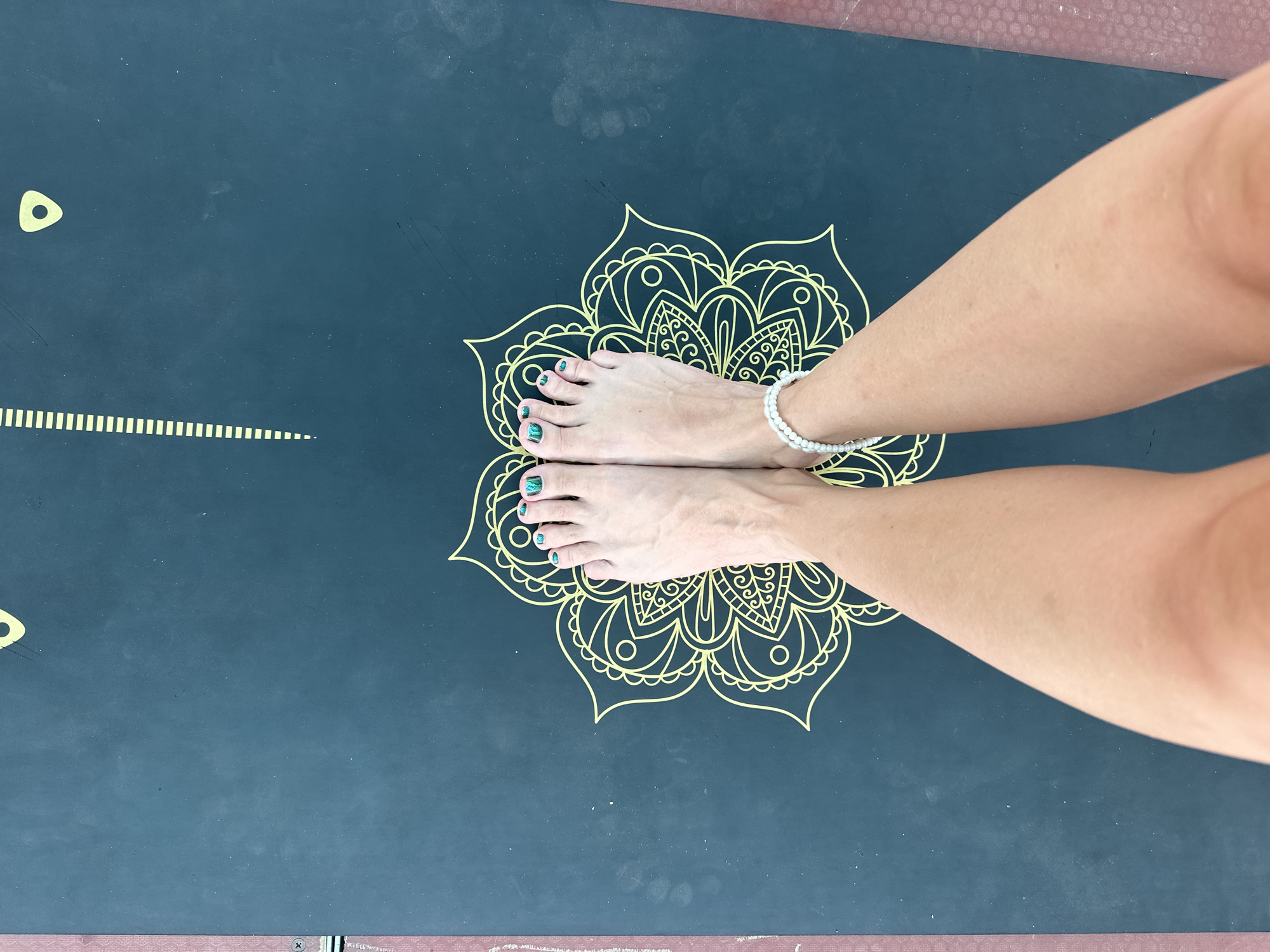 A top-down shot shows a pair of bare feet with green nail polish on a dark blue yoga mat with a gold mandala design. The feet are positioned in the center of the mandala. The individual is wearing a white beaded bracelet on their left ankle. The yoga mat has some scuff marks on its surface. The environment is indoors or outdoors, with no specific city details.