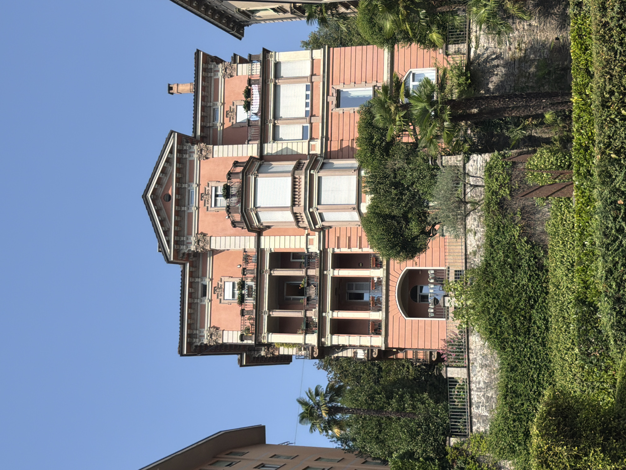 This is a medium shot of a coral-colored building in Lugano, Switzerland, taken on a clear, sunny day. The building, which appears to be an apartment building or hotel, features multiple balconies with railings and potted plants. The architectural style is ornate, with decorative molding and columns. A tall palm tree is visible to the left of the building, and other lush green vegetation covers the foreground and surrounds the building. The sky is a clear, bright blue. The image appears to be taken from a slightly elevated position, looking up at the building.