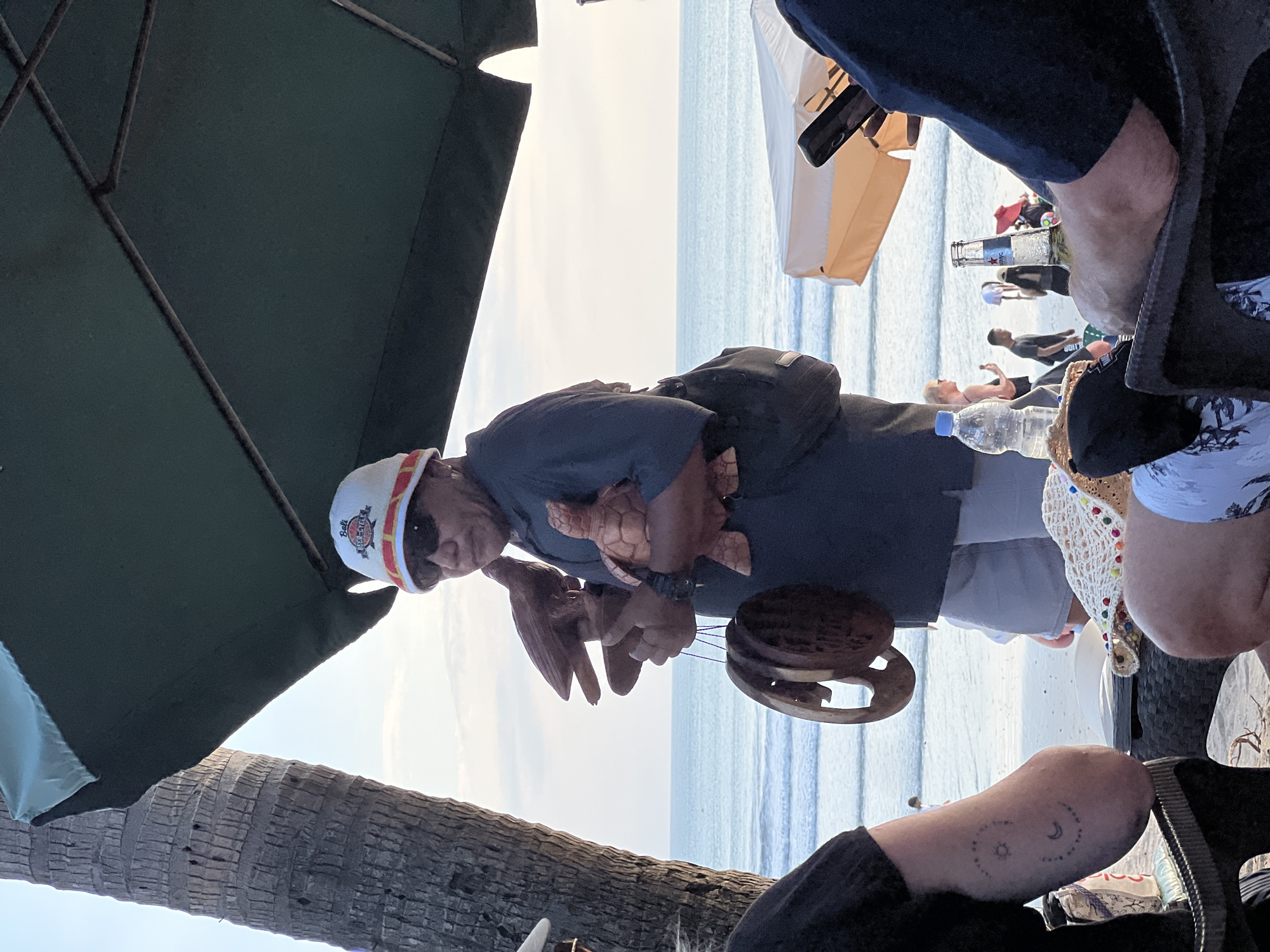 A man wearing a white hat with a red and yellow band, sunglasses, a dark blue shirt, and light-colored shorts stands on a sandy beach near the ocean. He holds several wooden carvings: a bird in his right hand and a turtle in his left arm. Below these, a circular wooden object hangs from a string. In the background, the ocean meets the sky under a cloudy sunset. Part of a palm tree is visible on the left, and a large dark green umbrella extends over the scene from the top. In the foreground, parts of people are visible sitting at tables, with a bottle of water and a beer bottle on one table. The man appears to be a vendor on the beach in Kuta, Indonesia, likely selling his crafts to tourists.