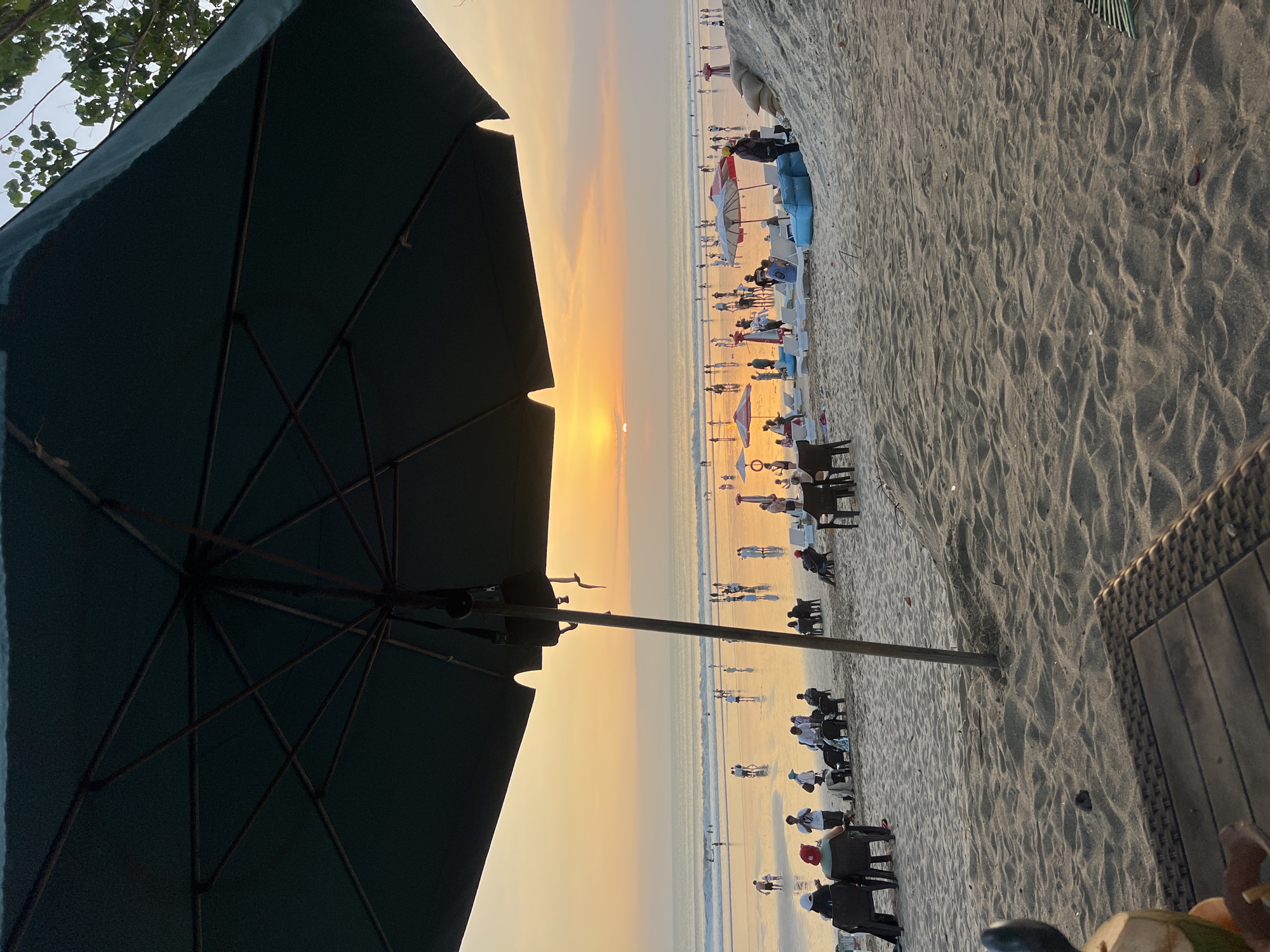 A wide-angle shot captures a bustling beach scene in Kuta, Indonesia, at sunset. The foreground is dominated by the textured sand and a dark green umbrella casting a shadow over the lower portion of the frame. To the left, a rattan table with a coconut drink is partially visible. In the mid-ground, several people are seated on chairs, some facing the ocean. Beyond them, the wet sand reflects the warm hues of the setting sun. The ocean stretches to the horizon, with gentle waves breaking on the shore and numerous people scattered along the water's edge, wading or strolling. Further out, small figures can be seen swimming. The sky is painted with vibrant orange and yellow tones, indicating the sun is low in the sky. Other umbrellas, lounge chairs, and beachgoers populate the middle and far grounds, contributing to the lively atmosphere of a popular tourist destination.