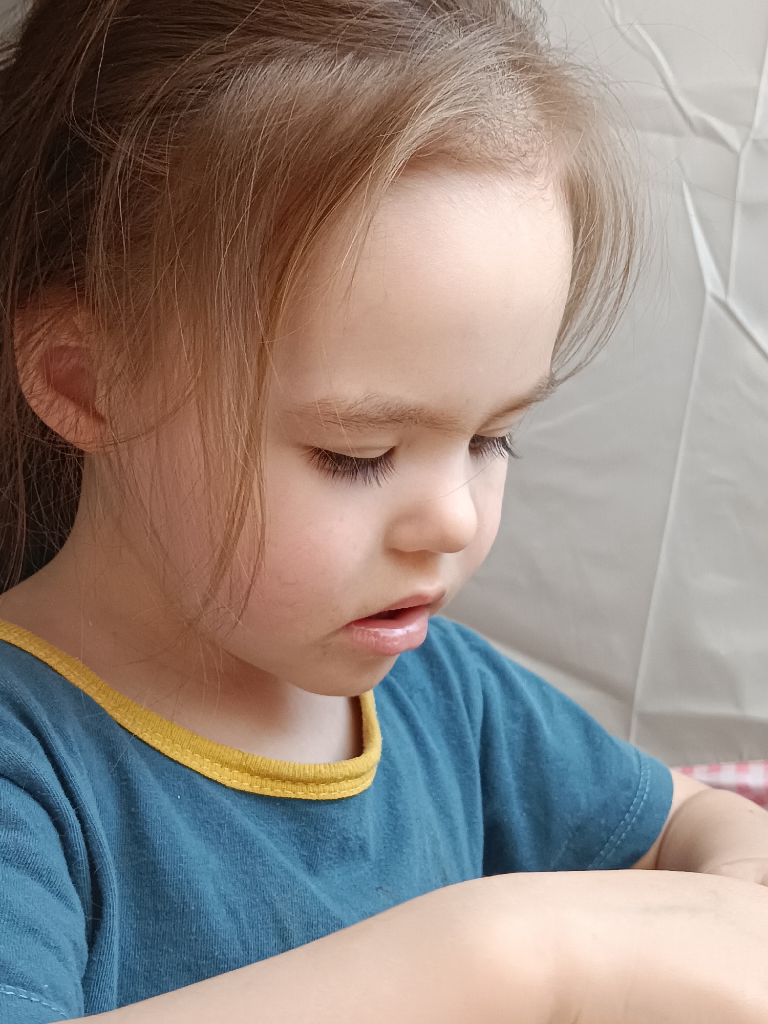 A close-up shot of a young girl with light brown hair pulled back. Her gaze is directed downward. She wears a teal t-shirt with a yellow collar. The background appears to be a wrinkled white surface. The location is not discernible from the image, and there is no indication it is in Noviy, Russia.