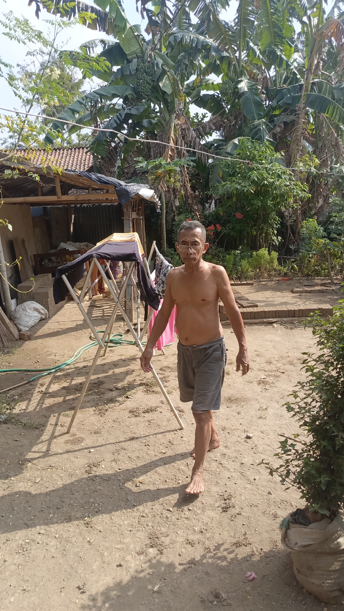An elderly shirtless man with short grey hair and glasses walks barefoot on a dirt path in front of a clothes drying rack in a yard. He is wearing grey shorts. The yard is filled with tropical plants, including banana trees, and there is a small building with a tin roof in the background. The scene appears to be in Kebumen, Indonesia, given the tropical vegetation and the architectural style of the building. The man is walking towards the viewer, and his expression is neutral. The lighting suggests it is a sunny day.