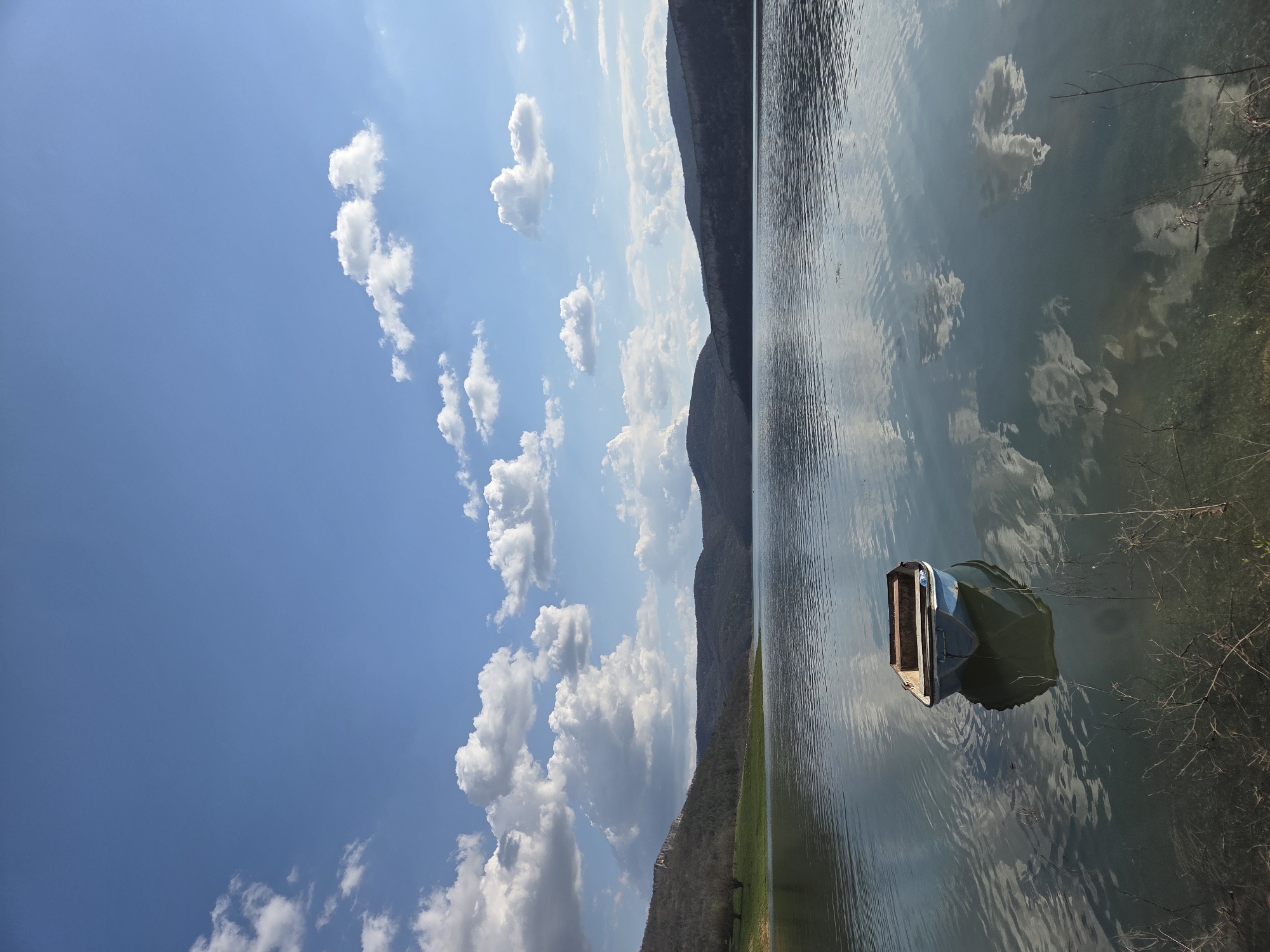 A landscape image rotated 90 degrees clockwise depicts a body of water with a boat, mountains, a sky filled with clouds, and hints of vegetation.
The sky occupies a significant portion of the left side of the image, exhibiting a bright blue expanse dotted with fluffy, white cumulus clouds.
The body of water dominates the right side, reflecting the sky and clouds, creating a mirror-like effect.
A small boat with an open back section floats on the water, near the shoreline. Its color appears a muted green or blue due to the lighting and reflection.
Mountains stretch across the middle of the image, separating the sky from the water.
The location could plausibly be near Debelets, Bulgaria, given the mountainous terrain and presence of a lake or river. The specific information connecting the image directly to Debelets is limited within the visual elements of the photograph.