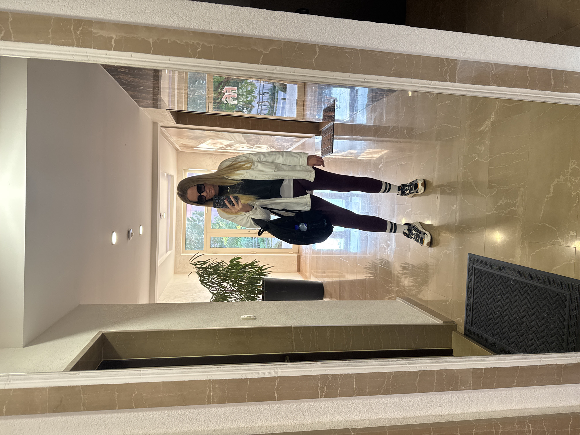A full-length mirror selfie captures a woman standing in an indoor hallway or lobby. The woman has long blonde hair and is wearing a white jacket over a black top with a visible "NIKE" logo, dark leggings or pants, and white sneakers with black and white striped socks. She is wearing dark sunglasses and holding a smartphone with a patterned case, using it to take the photo. A black backpack is visible on her shoulder.
The setting appears to be a well-lit interior space with light-colored, possibly marble or tiled, floors that have distinct veining patterns. Large windows in the background reveal outdoor greenery, including trees, and possibly parts of other buildings. A black doormat with a geometric border pattern is placed on the floor to the right. A potted plant with lush green leaves stands in a black planter near the windows. Recessed light fixtures are visible on a wall in the background. The bright natural light suggests it is daytime. Faint red text with a stylized character is visible in the reflection of an outdoor sign through the distant window, but it is unreadable. The location is in Budva, Montenegro.