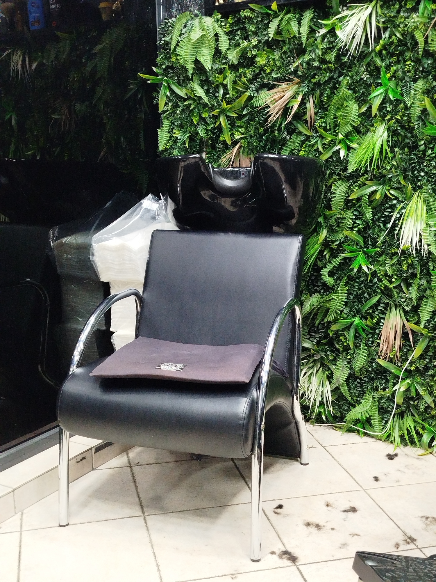 A black leather salon chair with chrome arms and legs sits in front of a backdrop of artificial foliage, possibly ferns and other greenery. The chair has a brown cloth pad on the seat. Above the chair is a black salon sink or basin. To the left, partially obscured, is a stack of white towels wrapped in plastic. The floor is tiled in light-colored squares, with clippings of hair scattered across it. The location is plausibly a hair salon in Varna, Bulgaria. No people are visible.