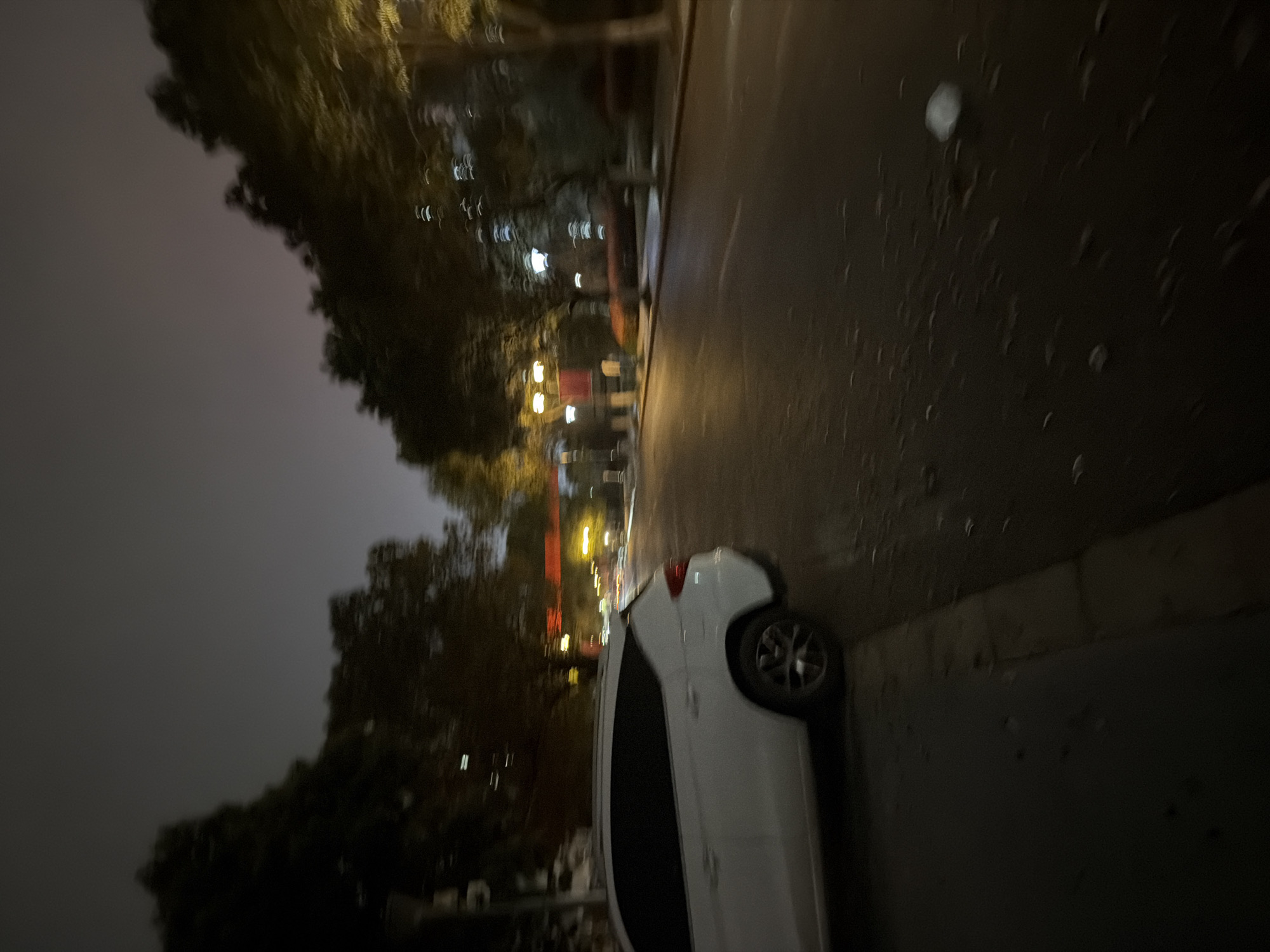 A blurry, dark, and slightly out-of-focus image shows a street scene at night. A white car is parked on the side of the road. Trees and streetlights are visible in the background, suggesting an urban environment. The location is likely Jiangning District, China. The image lacks sharpness and detail due to the low light conditions and possible motion blur.