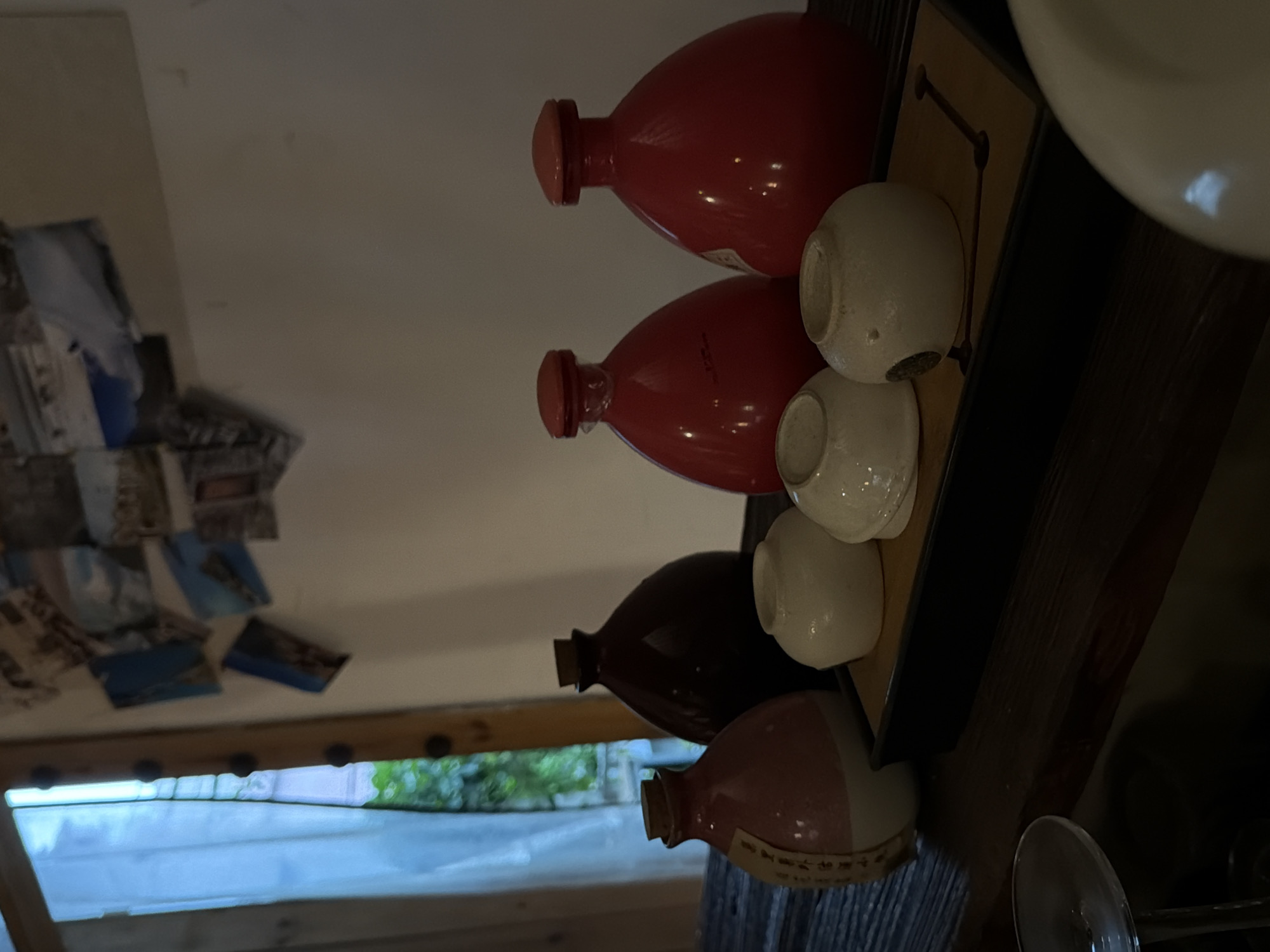 The image shows a close-up, eye-level view of a collection of ceramic bottles and cups arranged on a dark wooden surface, likely a shelf or table. The background features a window with sheer white curtains revealing a blurred view of greenery outside, and a wall adorned with several small photographs or postcards. The overall lighting is dim and warm, suggesting an indoor setting, possibly a home or a small shop in Longgang District, China.
In the foreground, a clear glass stemware is partially visible on the left, reflecting some light. The main subjects are the ceramic vessels. From left to right:
1. A pink and white ceramic bottle with a cork stopper and a label featuring what appears to be Asian script.
2. A dark brown, round ceramic bottle with a cork stopper.
3. Three small, off-white ceramic cups are placed on a dark rectangular tray with a lighter wooden insert that has carved patterns.
4. Two bright red, bulbous ceramic bottles with matching red stoppers stand behind the cups.
The photographs on the wall in the background are clustered and appear to depict landscapes or architectural scenes. The scene conveys a sense of curated display, possibly for sale or as decorative items. There are no people visible in the image.