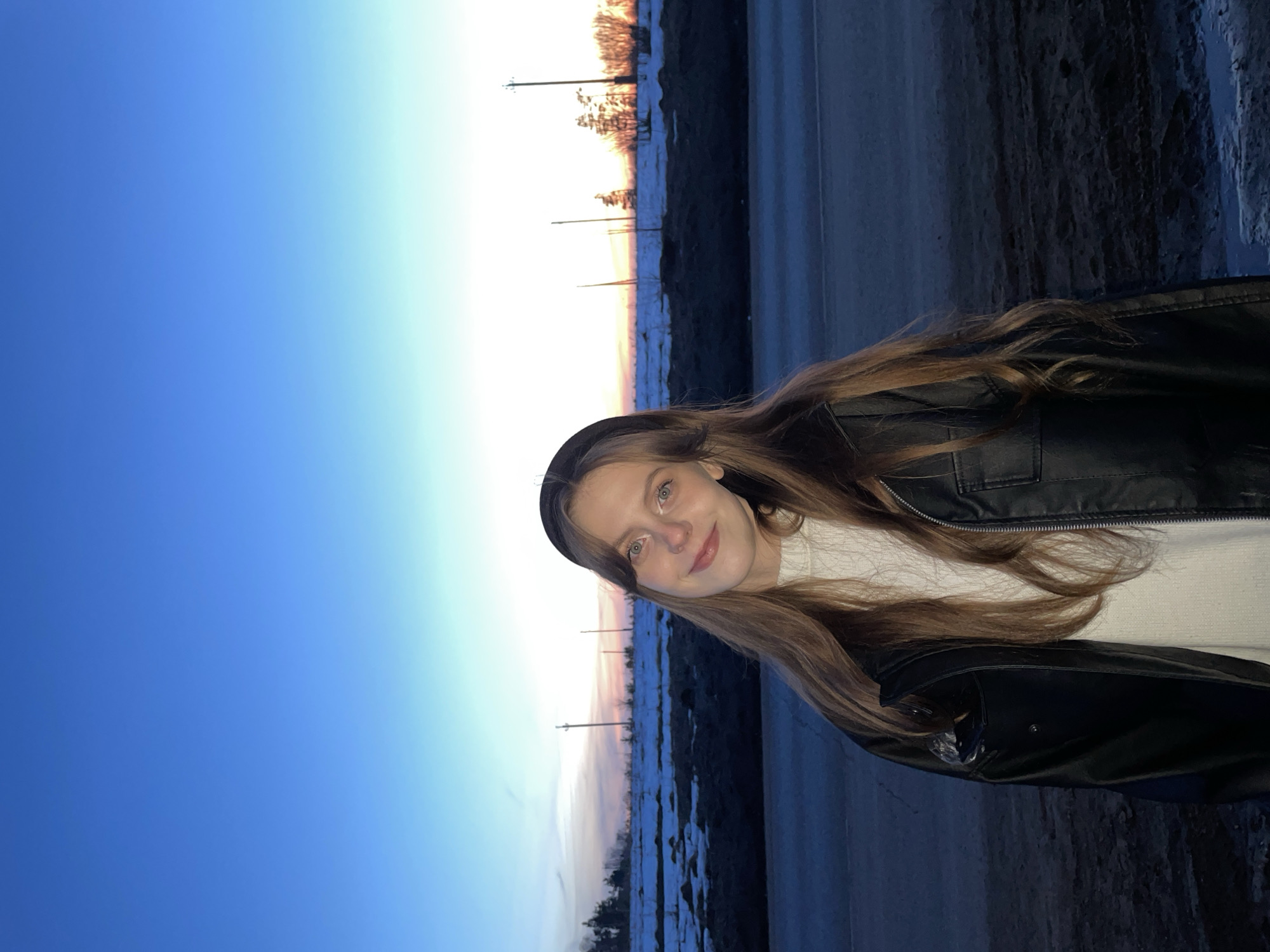 A young woman stands outdoors in the city of Фоки, Russia. She has long brown hair and wears a black leather jacket over a white sweater. Behind her is a road with snow-covered areas on the sides, leading towards a sunset. Utility poles are visible in the distance against the colorful sky. The time of day appears to be dusk or dawn.