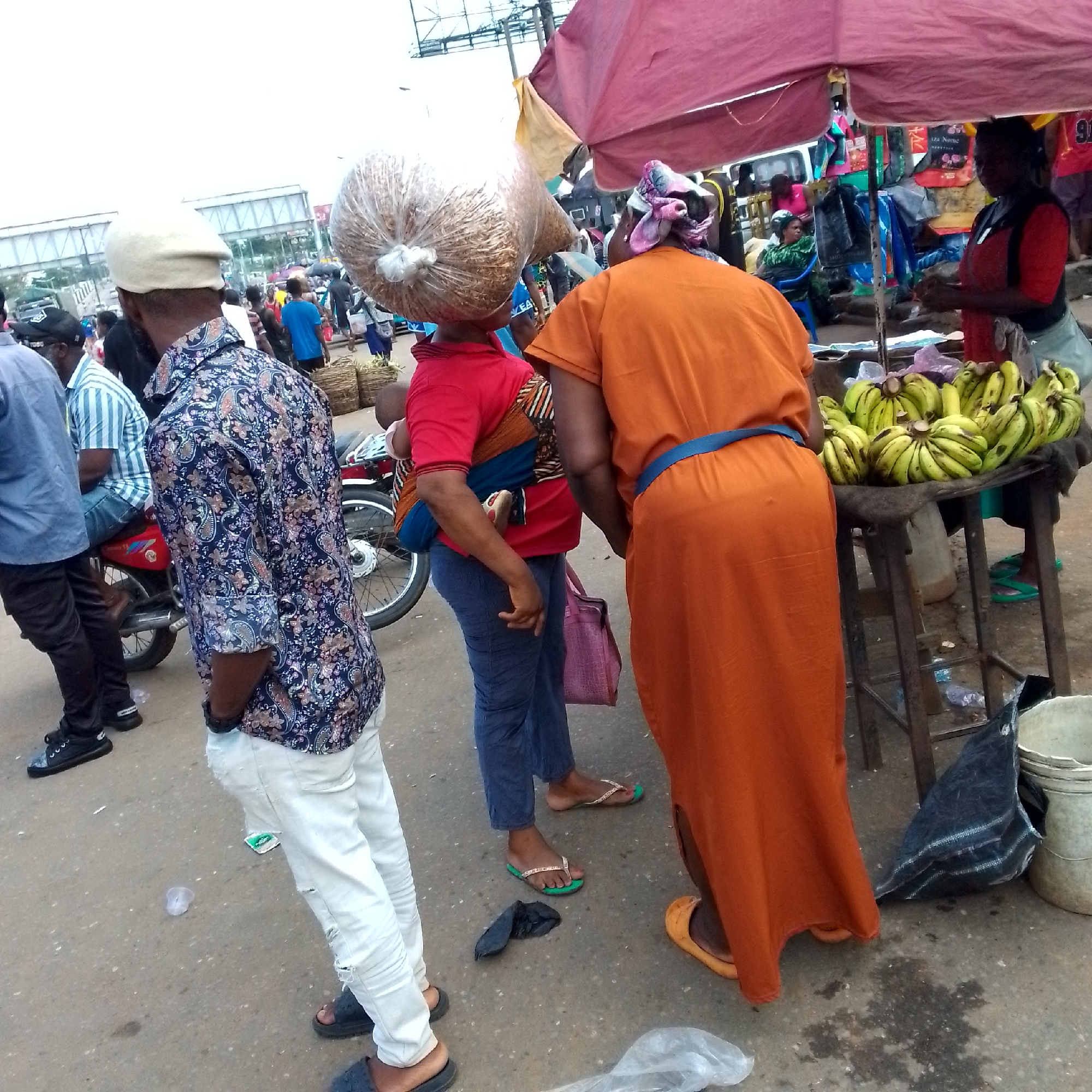 In Nkpor, Nigeria, a bustling street market scene unfolds. The foreground is dominated by the lower half of a person wearing ripped white trousers and dark sandals, facing away from the camera. To their left, another individual is captured from the waist up, dressed in a dark, patterned shirt. Their presence suggests a lively, crowded environment.
The midground reveals more of the market's activity. A woman, facing away, wears a vibrant orange dress with a blue belt. She stands near a stall laden with a bunch of ripe bananas. Beside her, another woman, in a red shirt and blue trousers, carries a baby wrapped in colorful fabric on her back. A large, clear plastic bag filled with what appears to be grain is balanced precariously on the head of a person in a red shirt. This distinctive image adds a touch of everyday resourcefulness to the scene.
The background shows a glimpse of the urban setting with more people, stalls, and a motorcycle. The ground is paved and somewhat littered, typical of a busy market area. A red umbrella shades a portion of the market. The overall impression is one of a dynamic and active marketplace, alive with the commerce and daily routines of Nkpor.