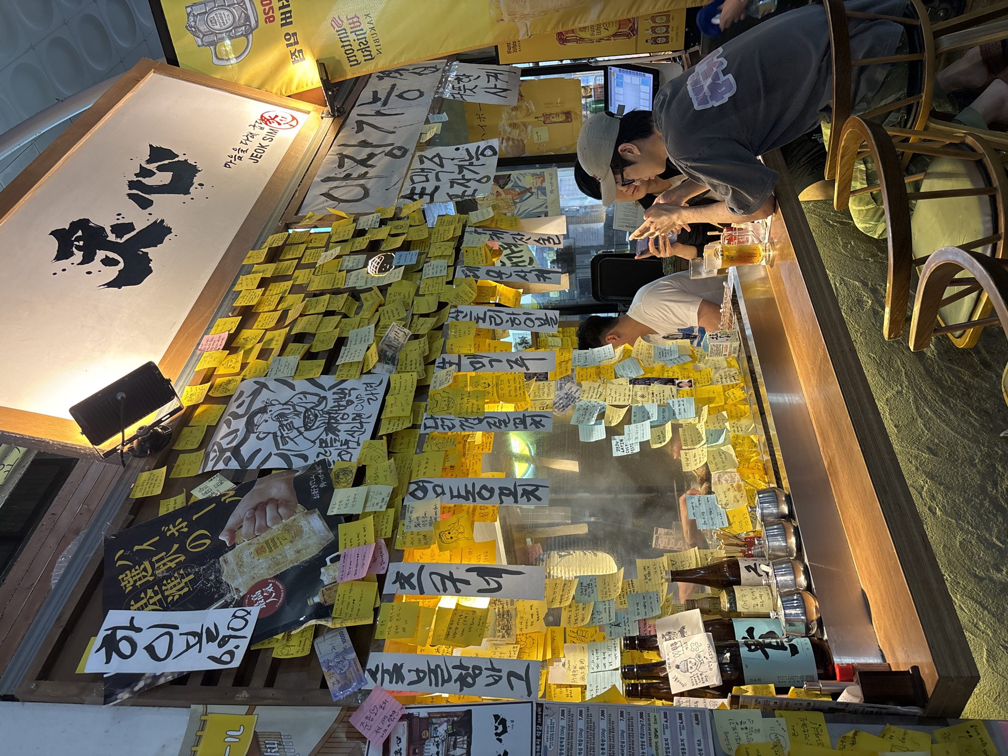 This media file captures a lively scene inside a bar or restaurant, likely in Seogwipo-si, South Korea, given the provided location context. The primary subjects are several individuals, predominantly young men, seated at a wooden bar. They appear engaged in conversation and interaction, with some looking at their phones. The atmosphere seems relaxed and convivial, suggesting a casual outing.
The setting is characterized by a wall densely covered in colorful sticky notes, many with handwritten messages and drawings in Korean and possibly Japanese. This creates a vibrant and personalized decor. Behind the bar, shelves are stocked with various bottles, including sake and beer, indicating the establishment's offerings. A prominent sign with Korean calligraphy, "마음을 다해 굽단다 JEOK SIM," which translates to "Grilling with all my heart JEOK SIM," suggests the name of the establishment and its focus on grilled food. Other visible text includes drink names like "하이볼" (highball) and "日本酒" (sake), as well as menu prices and personal notes left by patrons.
A spotlight illuminates a section of the wall adorned with a large graphic advertisement, possibly for food or drink, featuring a hand pouring a drink. The lighting suggests it could be evening or indoors, with a warm, inviting ambiance. The overall impression is one of a popular, community-oriented spot where customers feel comfortable leaving their mark.